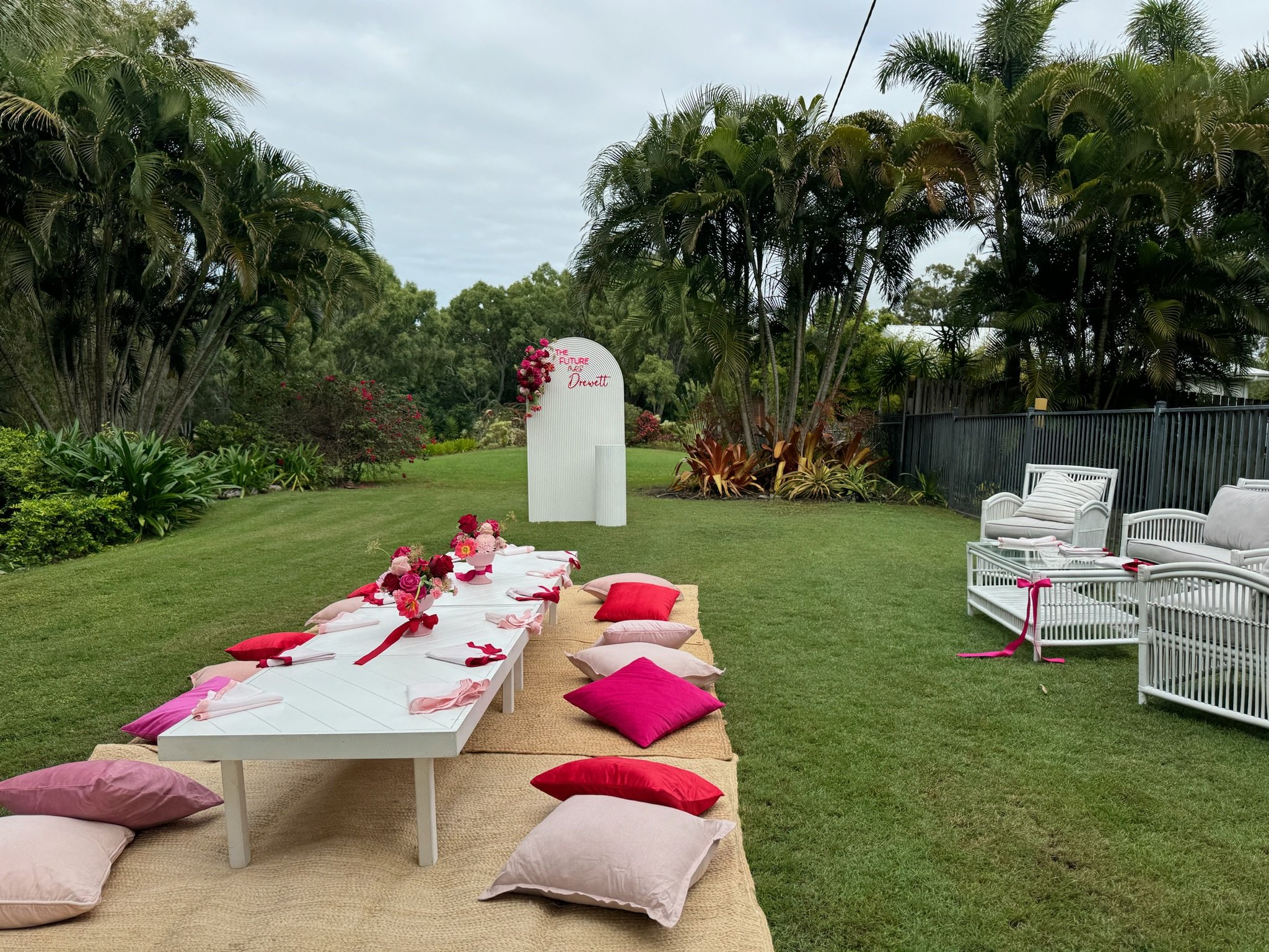 Outdoor garden event setup with low tables, cushions, pillows, and floral decorations, featuring a white backdrop with pink lettering in the background.
