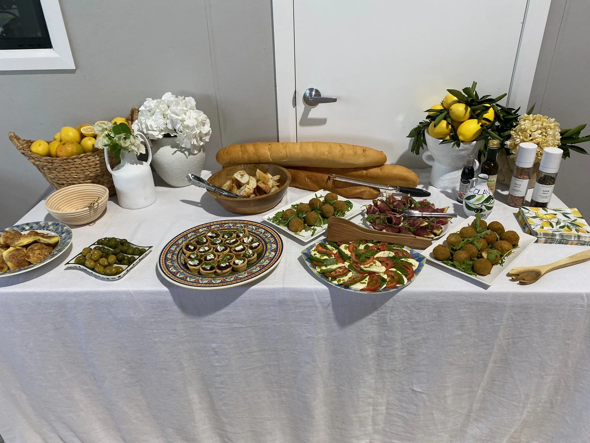 A table set with bread, salads, appetizers, and condiments, decorated with lemon and hydrangea arrangements in a bright room.