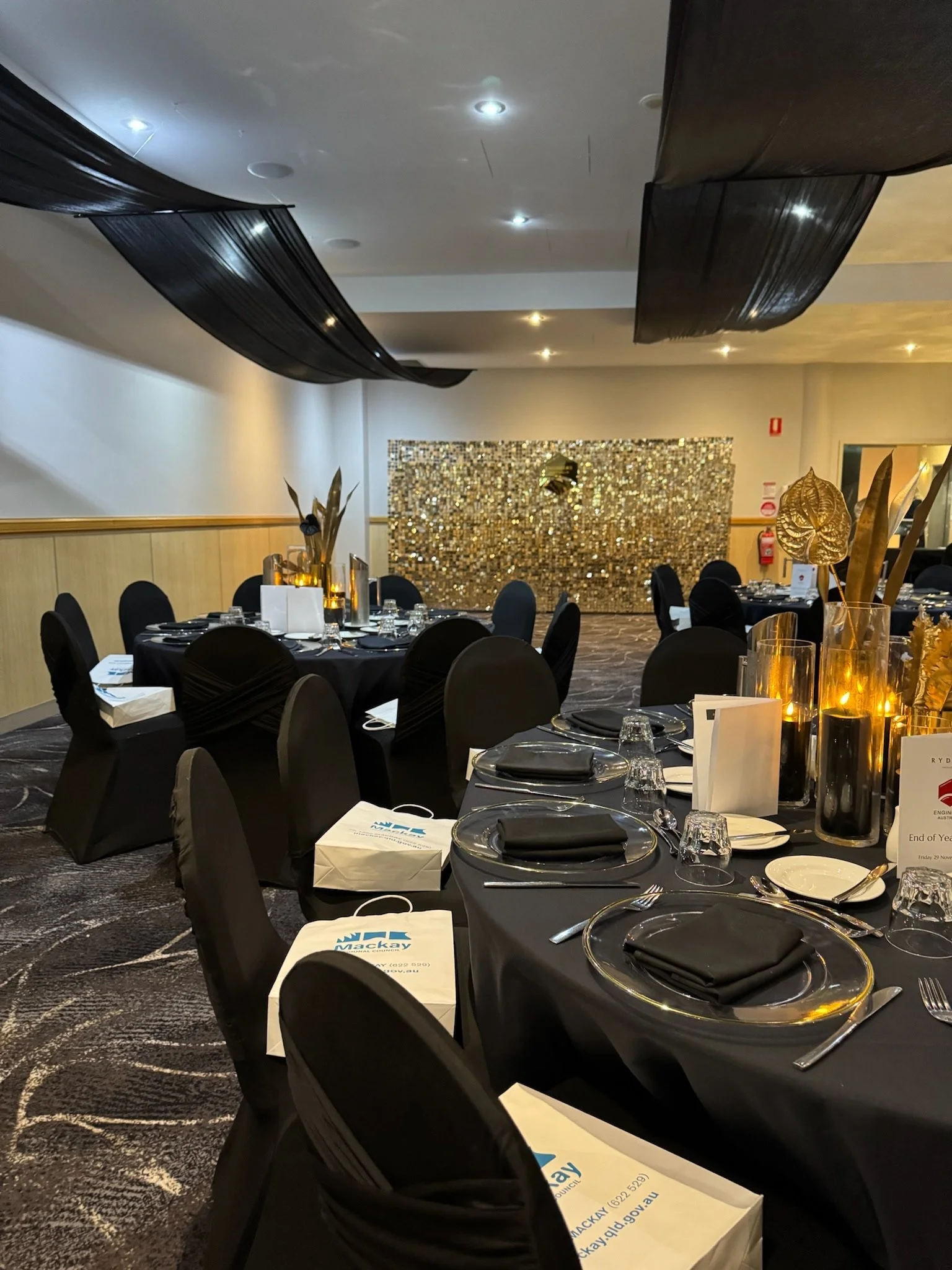 Event banquet hall with round tables covered in black tablecloths, decorated with gold and black centerpieces, set with plates, glasses, and cutlery, and featuring a golden sequin backdrop on the far wall.