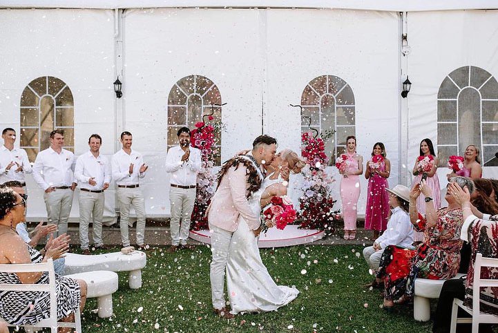 Bride and groom dancing at their wedding reception, with guests clapping and watching, in a decorated outdoor venue with pink and red floral arrangements.