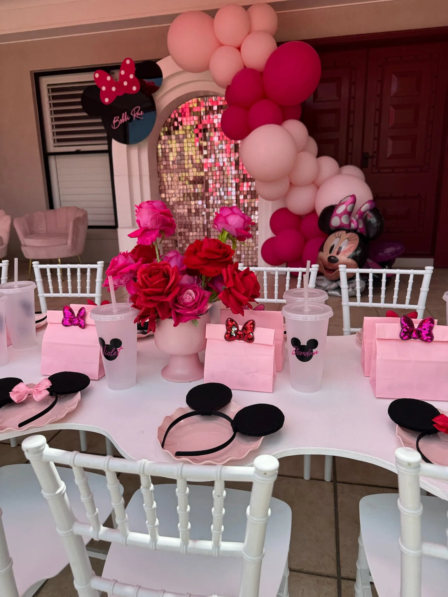 Party table decorated with pink and black Minnie Mouse themed items, including Minnie Mouse ears headbands, a pink flower centerpiece, pink and black balloons, and Minnie Mouse lanterns, in a pink and white theme for a birthday celebration.