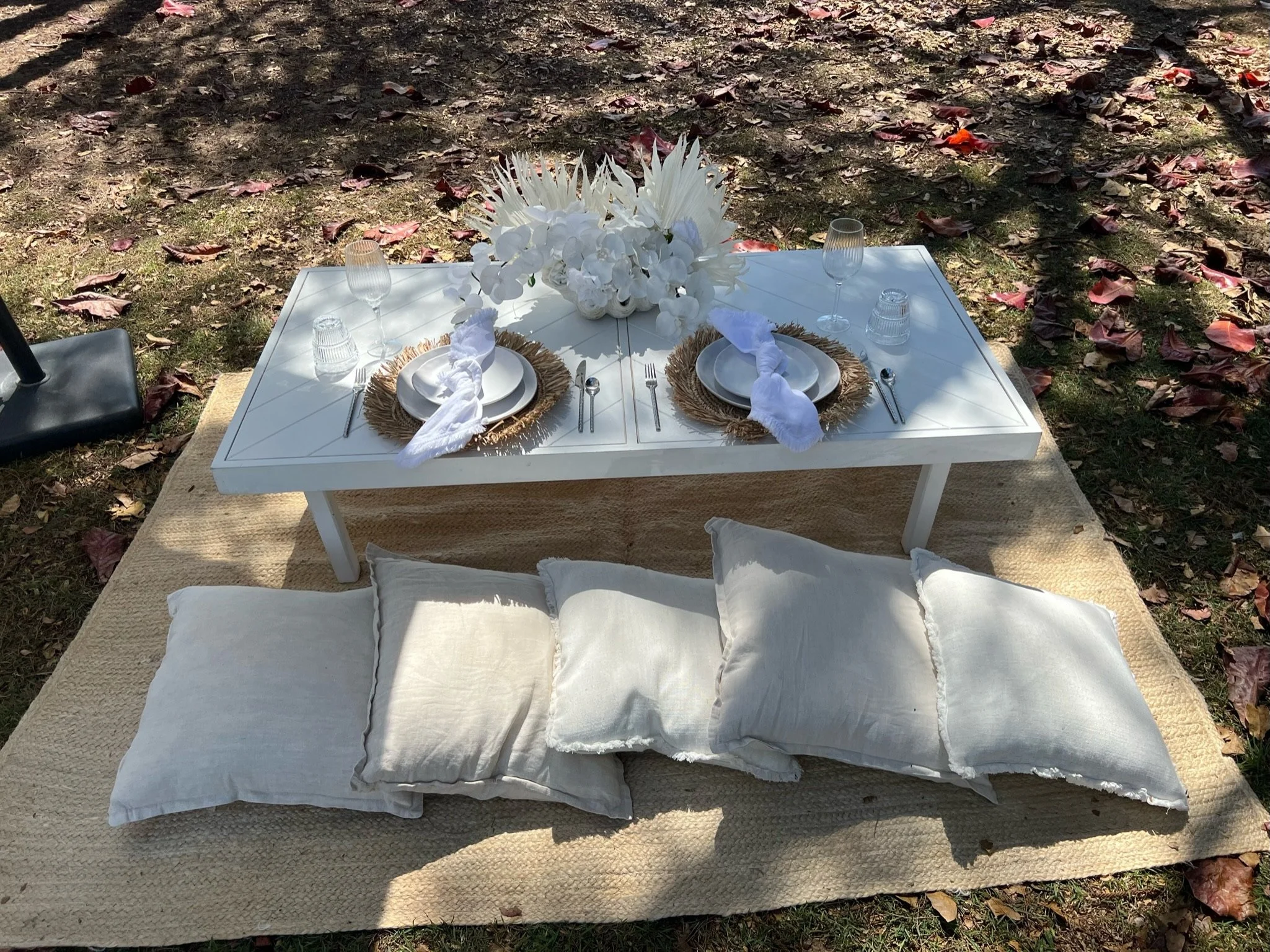 A low white table set for an outdoor meal with two place settings on woven placemats, white plates, linen napkins, silverware, with a floral centerpiece and four glasses, on a rug amidst fallen autumn leaves.