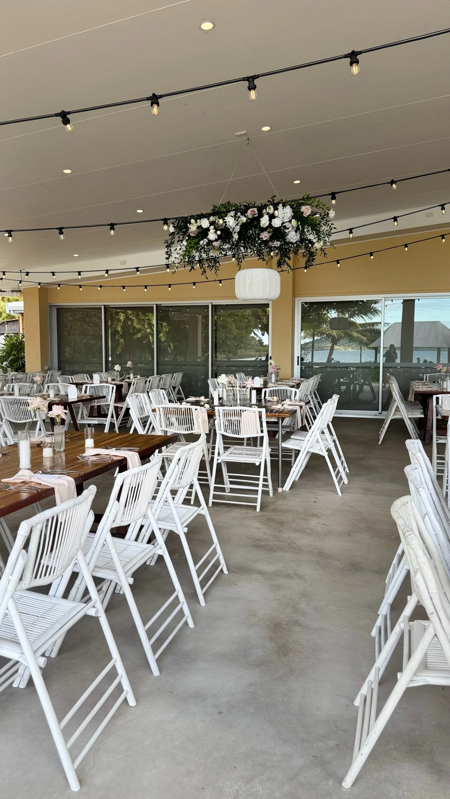 A decorated outdoor dining area with wooden tables, white chairs, and string lights hanging from the ceiling. There is a floral arrangement hanging from the ceiling and a white lantern light. Large glass windows show a view of trees and water outside