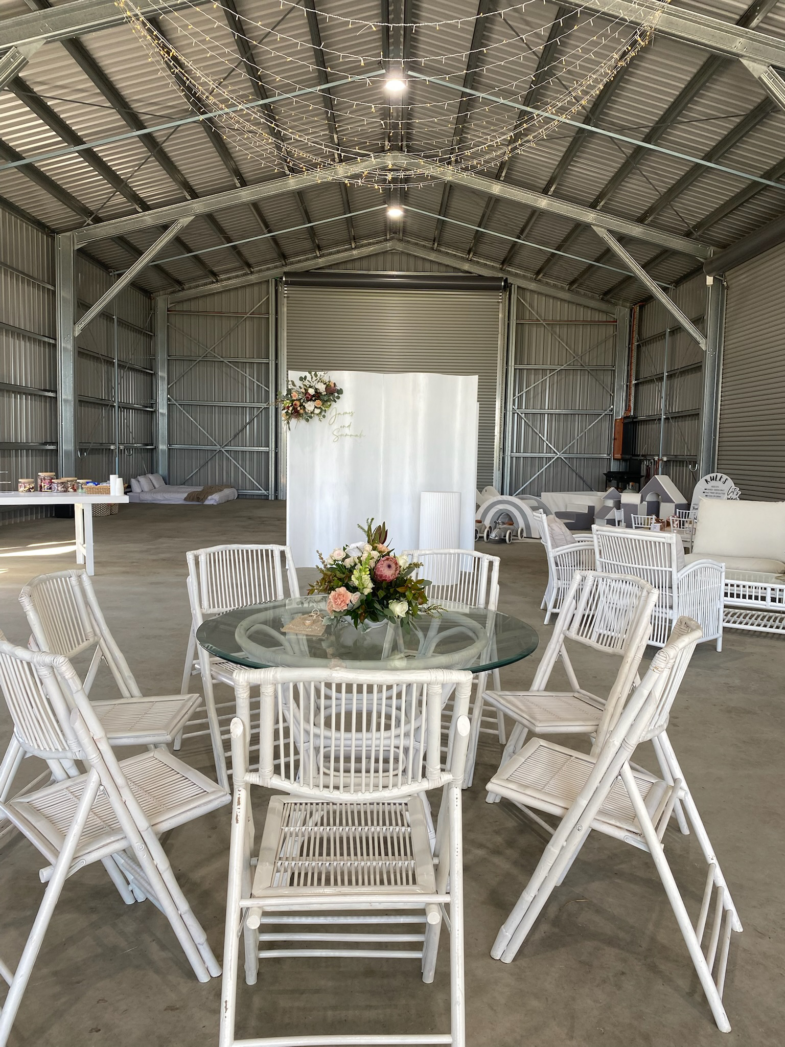 Indoor event space with a round glass table and white chairs, floral centerpiece, decorated backdrop with flowers and writing, and string lights hanging from the ceiling.