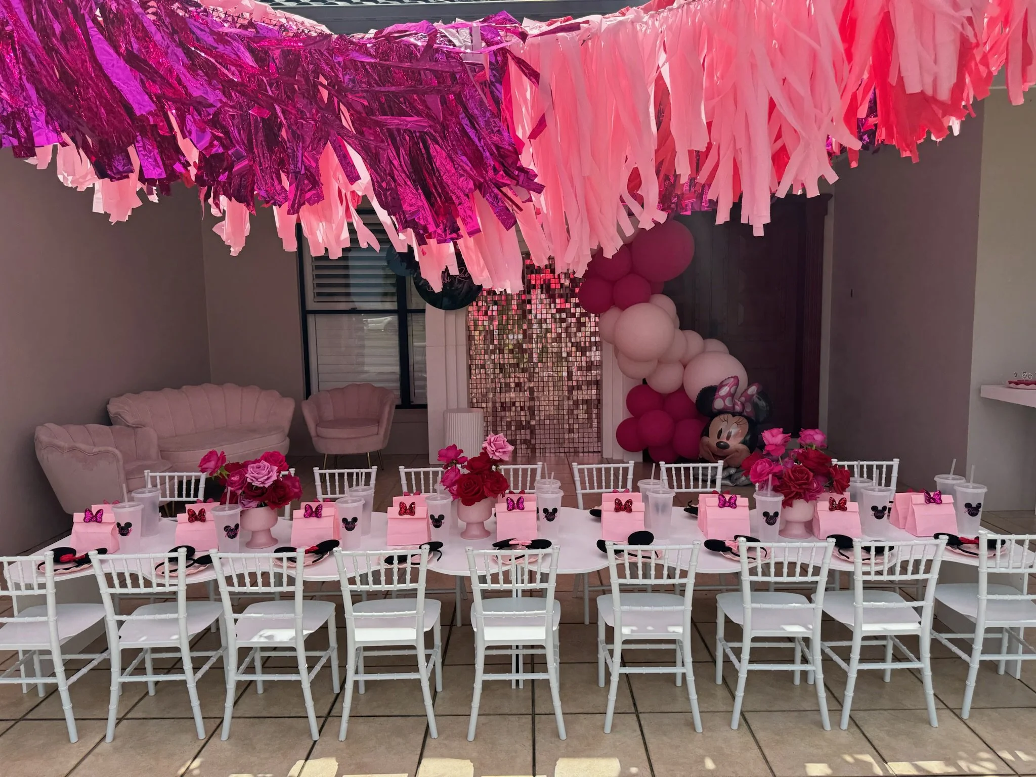Decorated party area with a long table set for a celebration, featuring pink flowers, pink boxes, Minnie Mouse themed cups, and black bows. Overhead, pink, purple, and rose gold metallic streamers hang. In the background, pink and white balloon arran