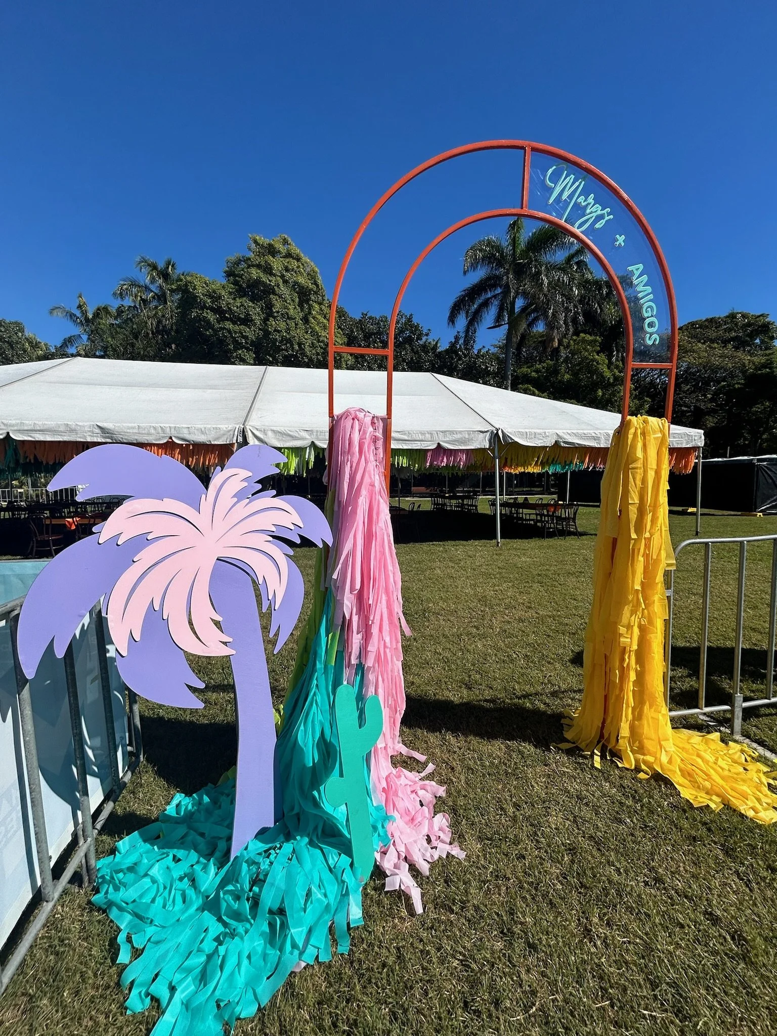 Decorative tropical-themed arch with pink, blue, and yellow fabric streamers and cutouts of palm trees and cacti, set up outdoors in front of a white tent and lush green trees under a clear blue sky.
