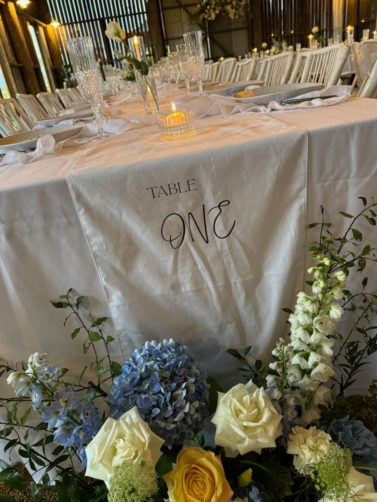 Wedding reception table with white tablecloth, floral arrangements, glassware, and candles, marked as 'Table One'.