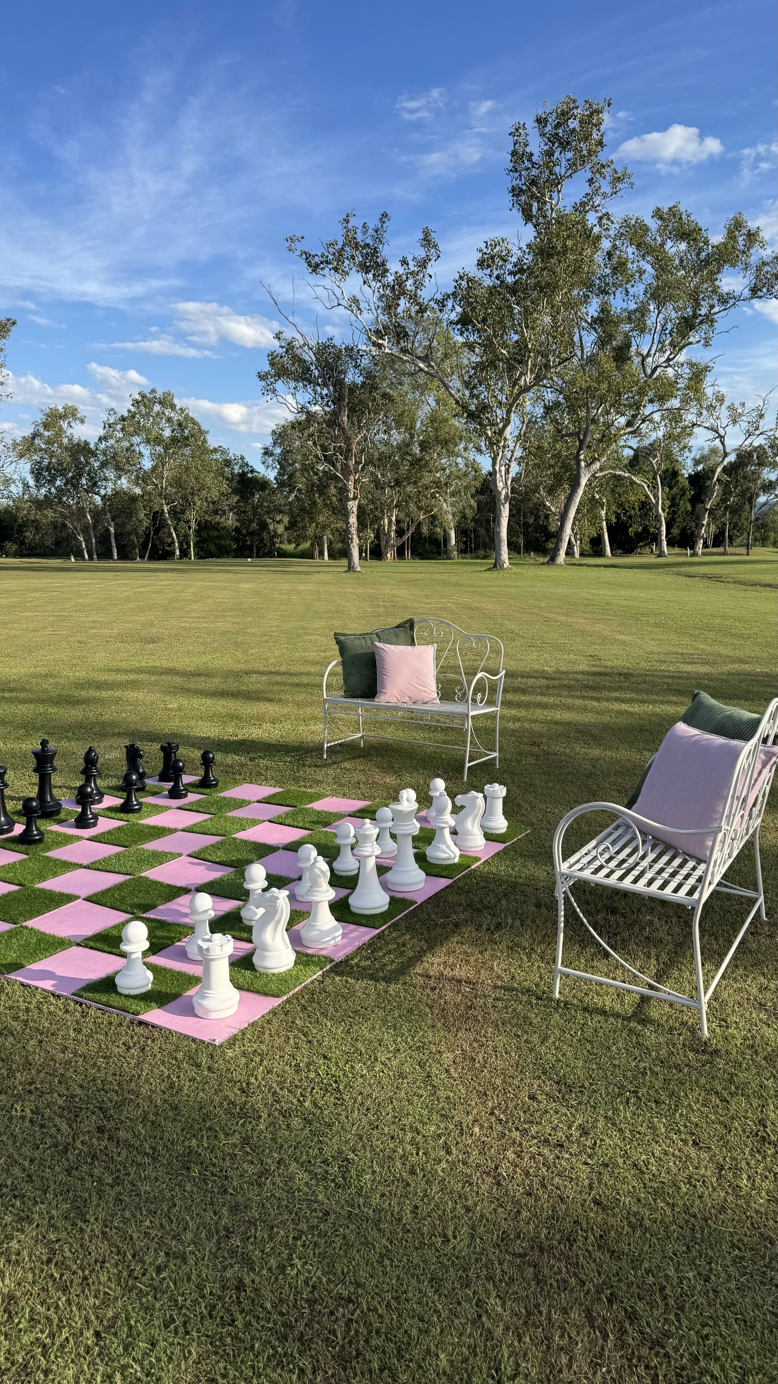 An outdoor scene with a large red and black chessboard on the grass, surrounded by white metal chairs and a white bench with pillows. Tall trees and a blue sky with clouds are in the background.