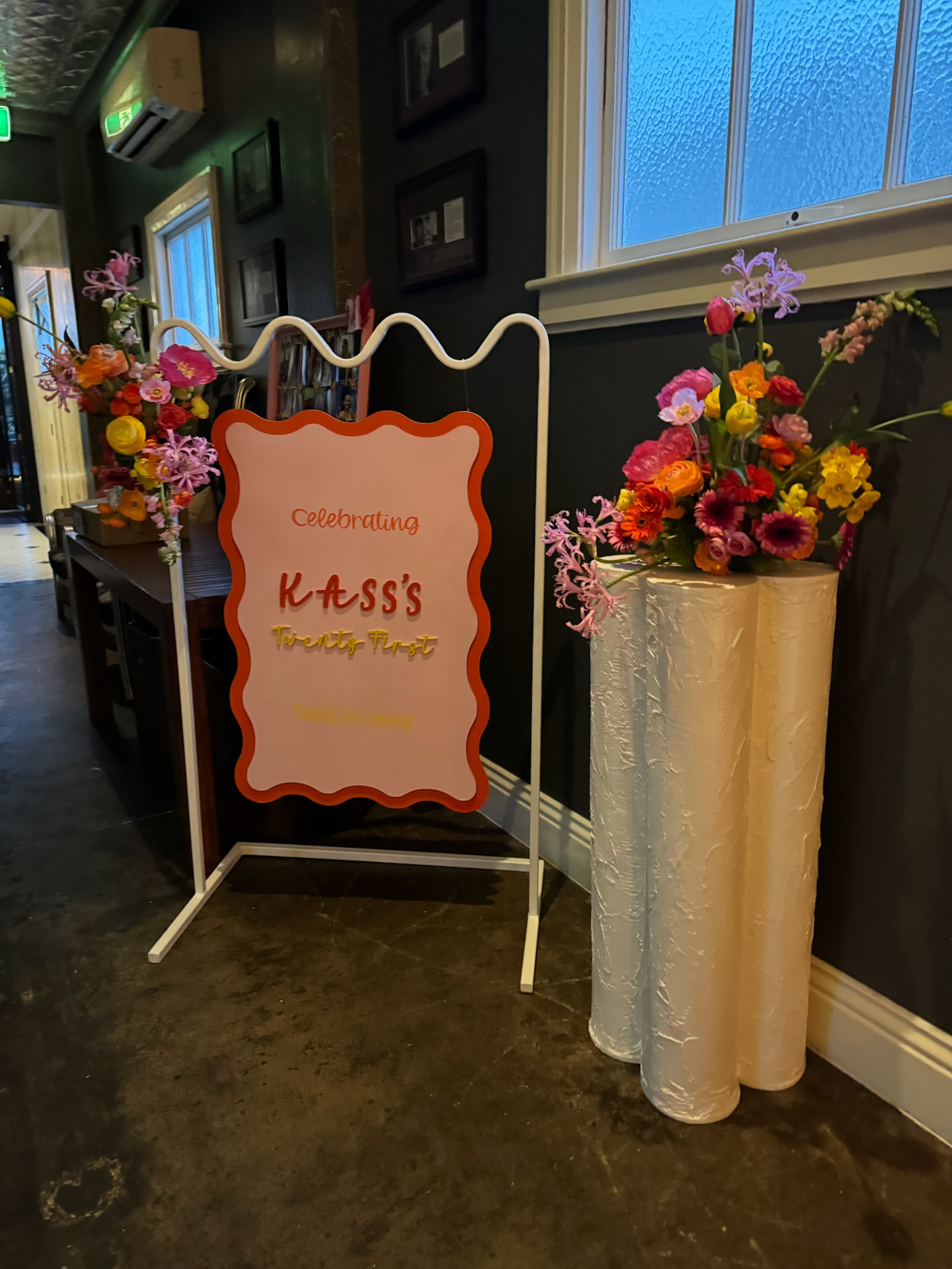 Celebration sign for Kass's twenty-first birthday, with floral arrangements on tall white cylinders.