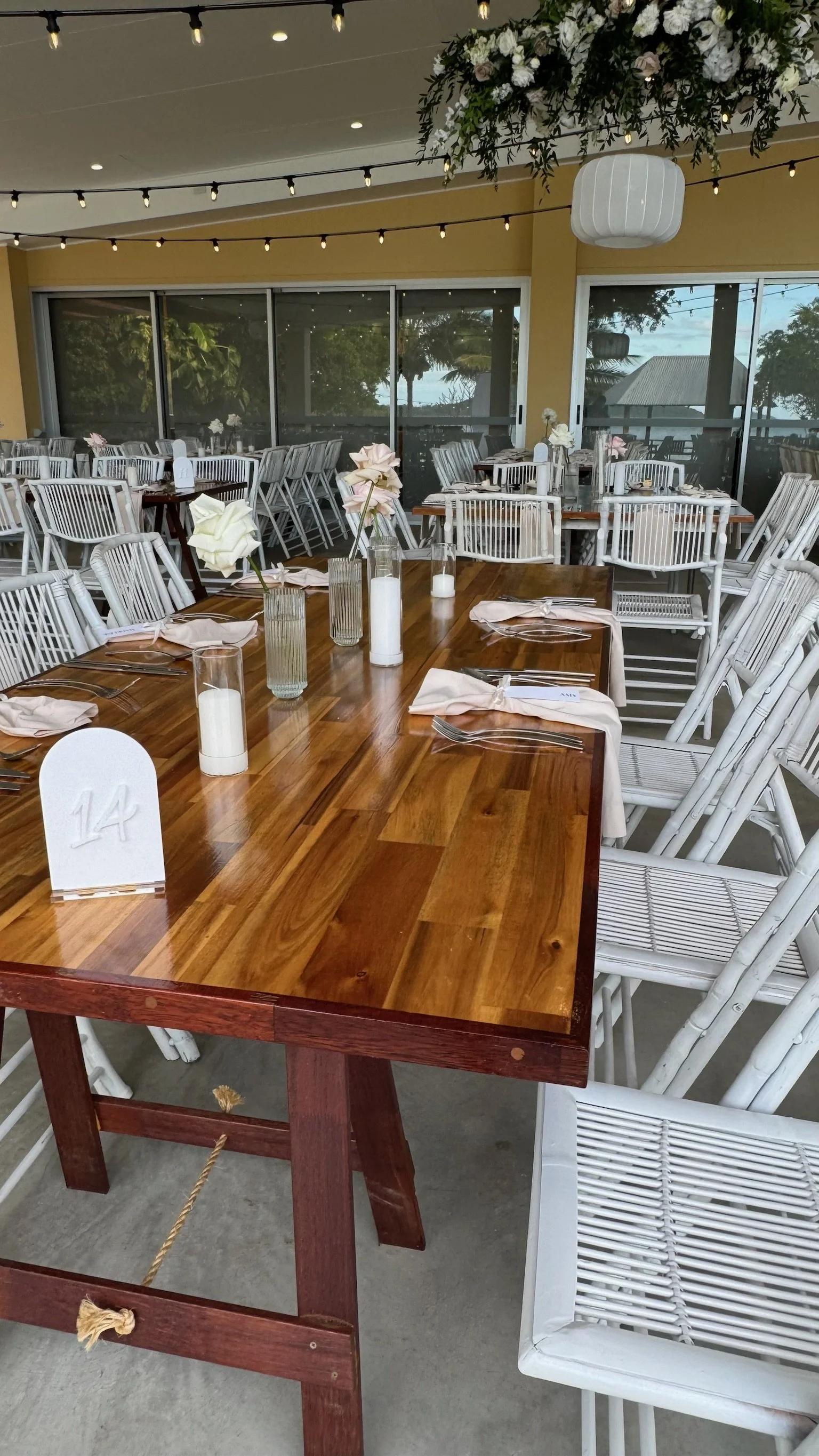 Decorated banquet table with white chairs, tableware, candles, and flower centerpieces in a bright dining area with large windows and string lights.