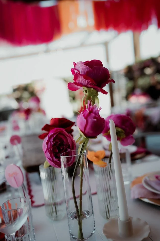 Decorative pink flowers in a glass vase on an elegant table setting with candles and glassware, possibly at a celebration or event.