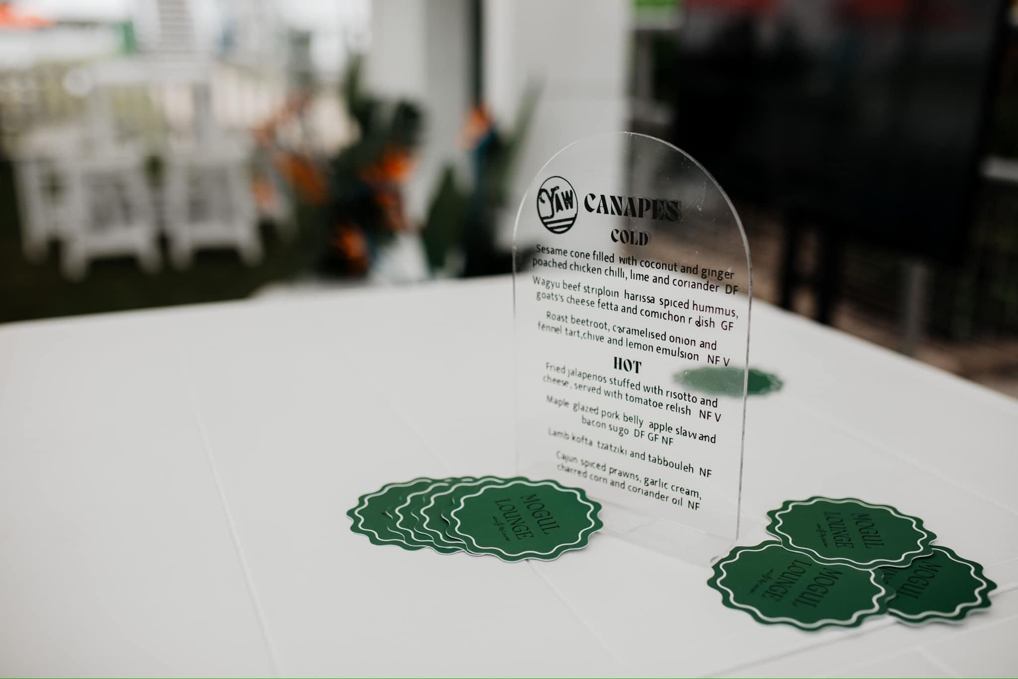 A clear acrylic menu card with black text and green coaster cards on a white surface. The menu features cold and hot dishes such as sesame cone, Wagyu beef striploin, roast beetroot, falafel tart, and Cajun spiced prawns.