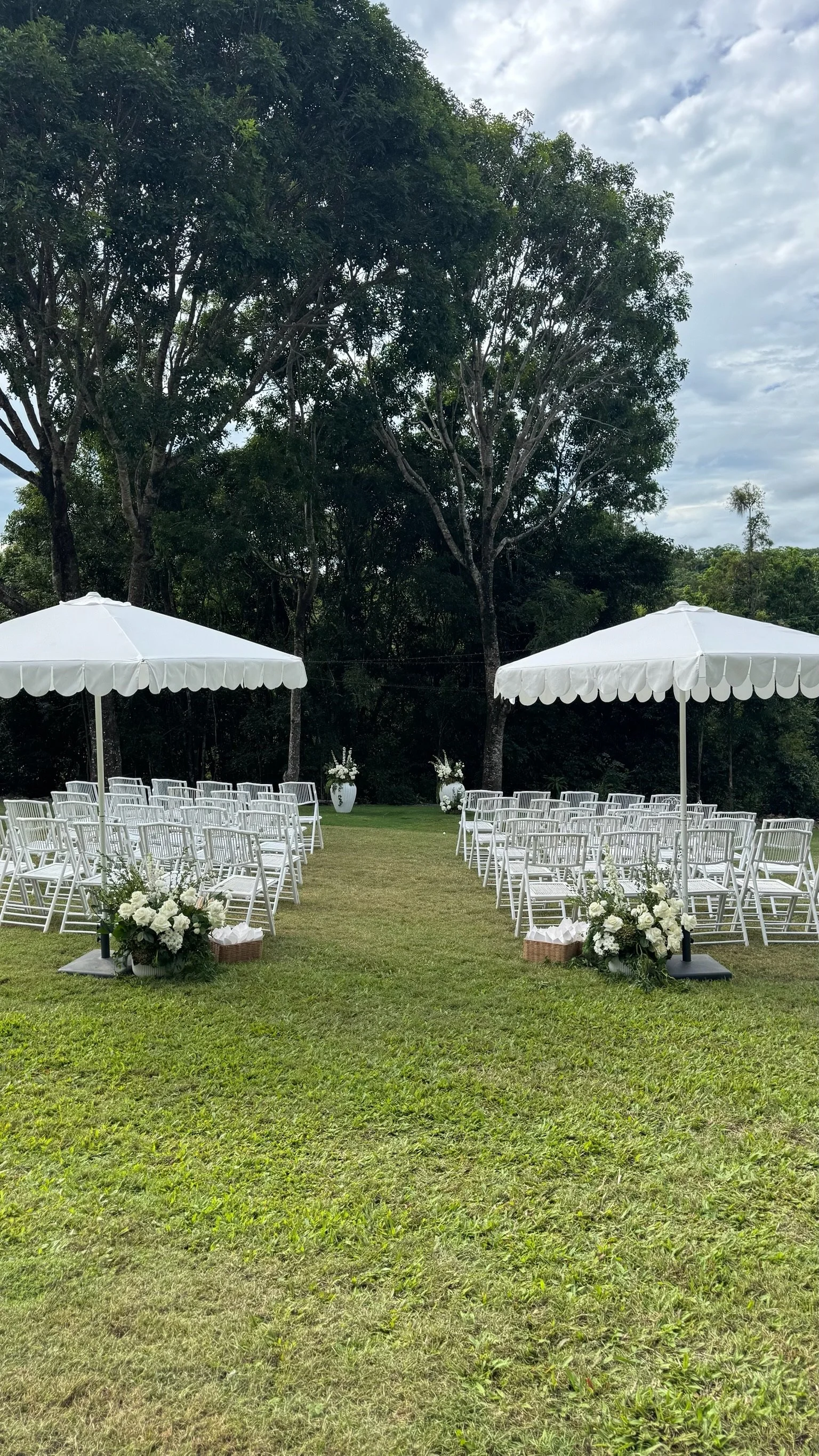 Outdoor wedding setup with white chairs, white umbrellas, floral arrangements, and large trees in the background.