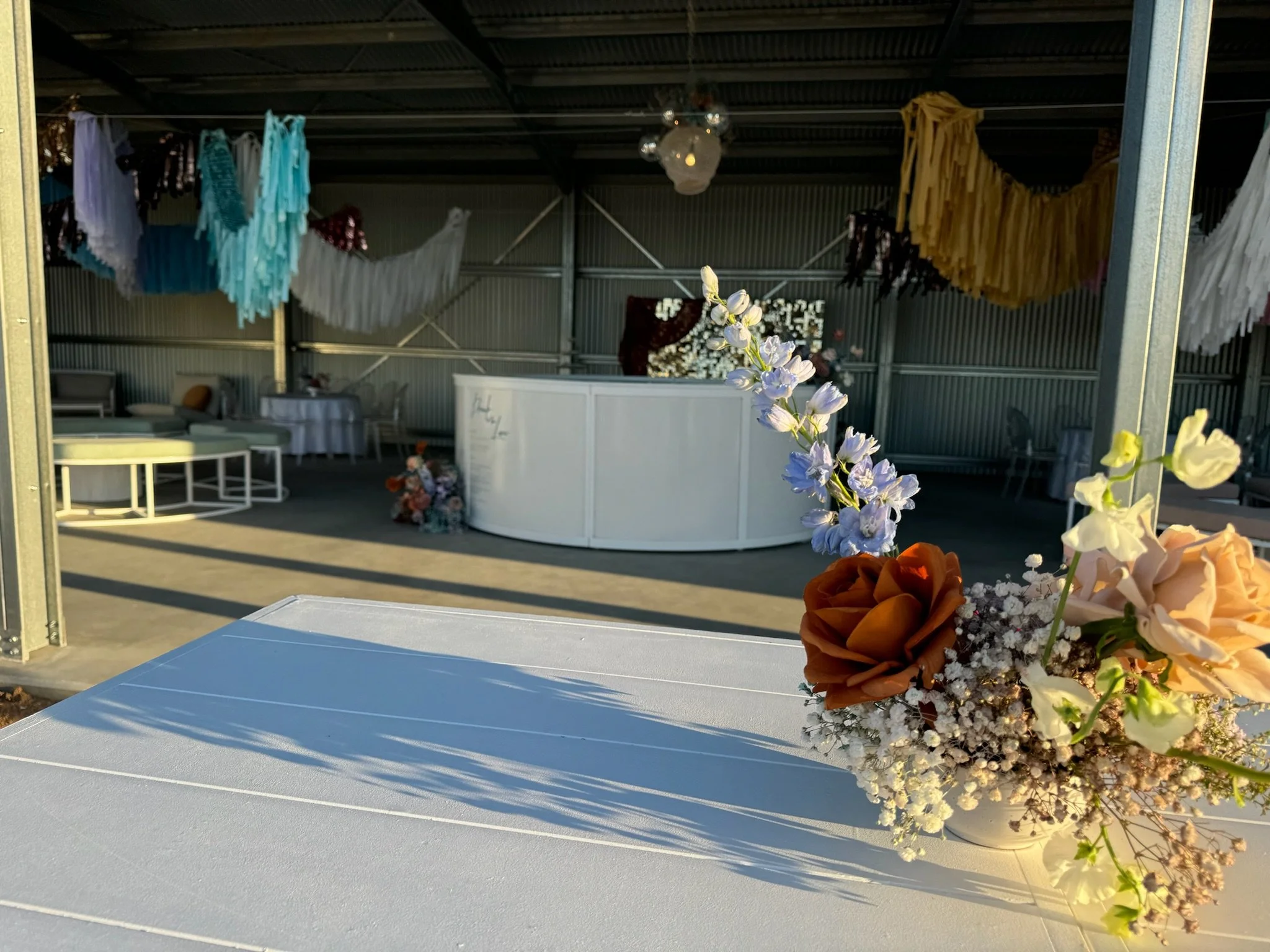 Close-up of a floral arrangement with brown, white, and light purple flowers on a white table, with event decorations and seating in the background.