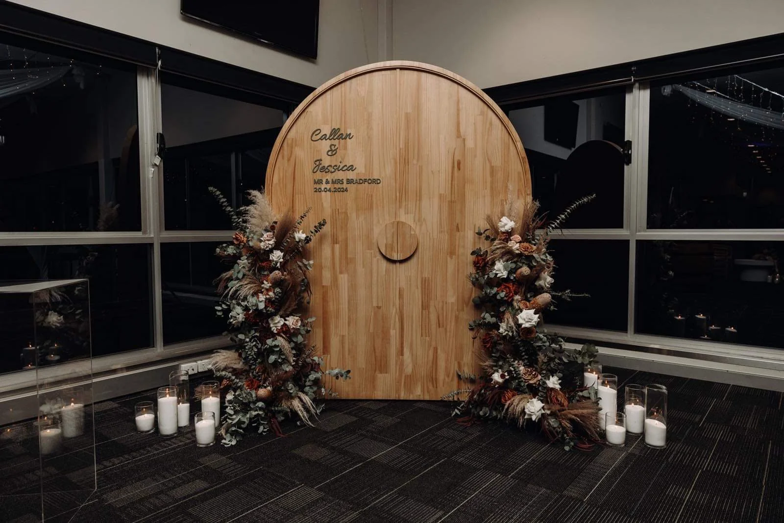 Wedding backdrop featuring a tall wooden arch with floral arrangements on both sides and candles at the base. The arch has text including names 'Callan & Jessica' and a date '20.04.2024'.