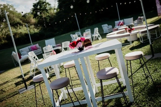 Outdoor event setup with white tables, pink and white chairs, and floral centerpieces, under string lights.
