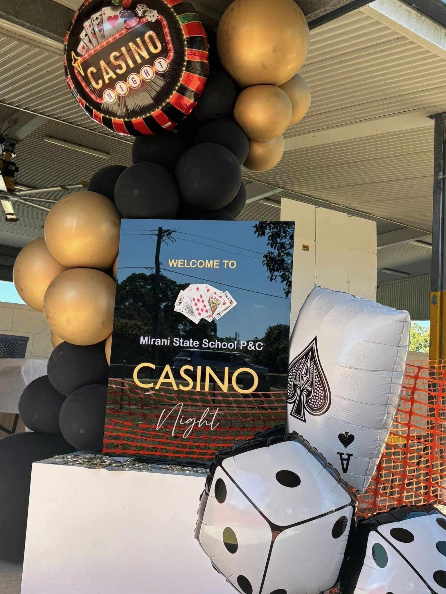 Decorative casino-themed balloons and a sign welcoming attendees to a casino night event at Mirani State School P&C. The balloons include a casino sign, large gold and black spheres, and dice-shaped balloons.