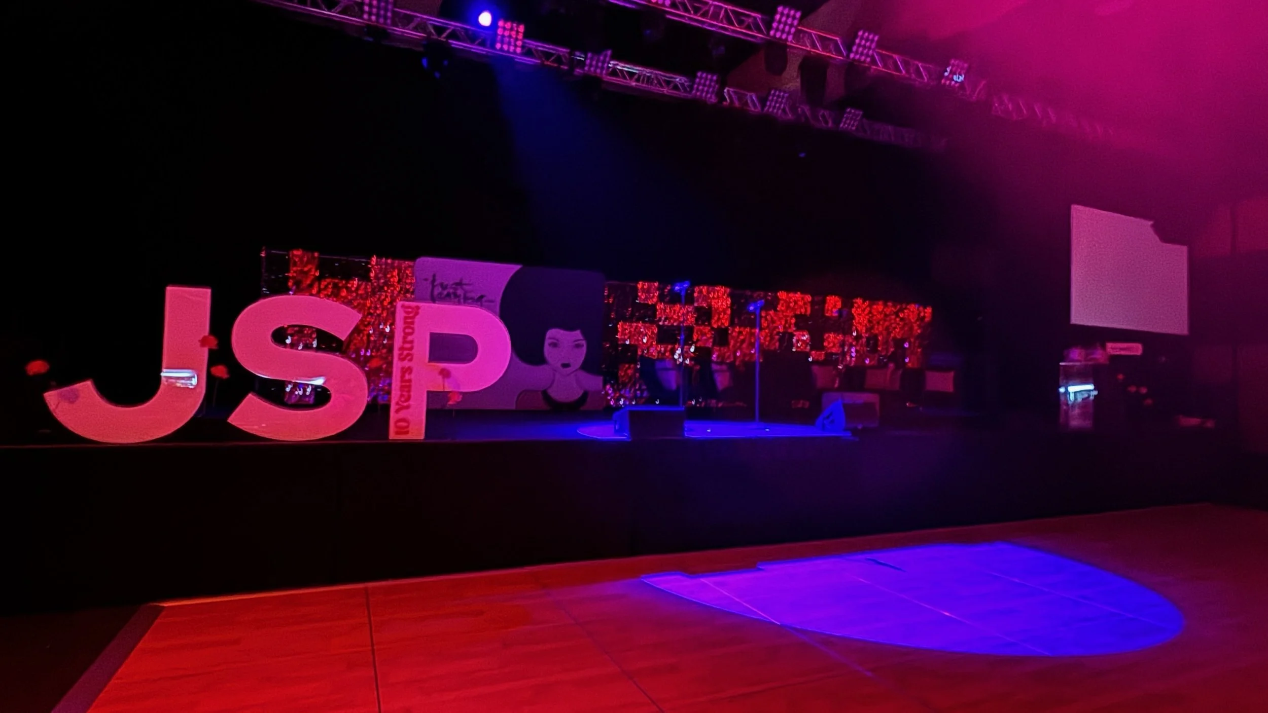 Stage decorated with large illuminated letters spelling 'JSP' and a red glitter backdrop, set for a celebration or event, under purple and blue lighting, with empty chairs and screens visible.