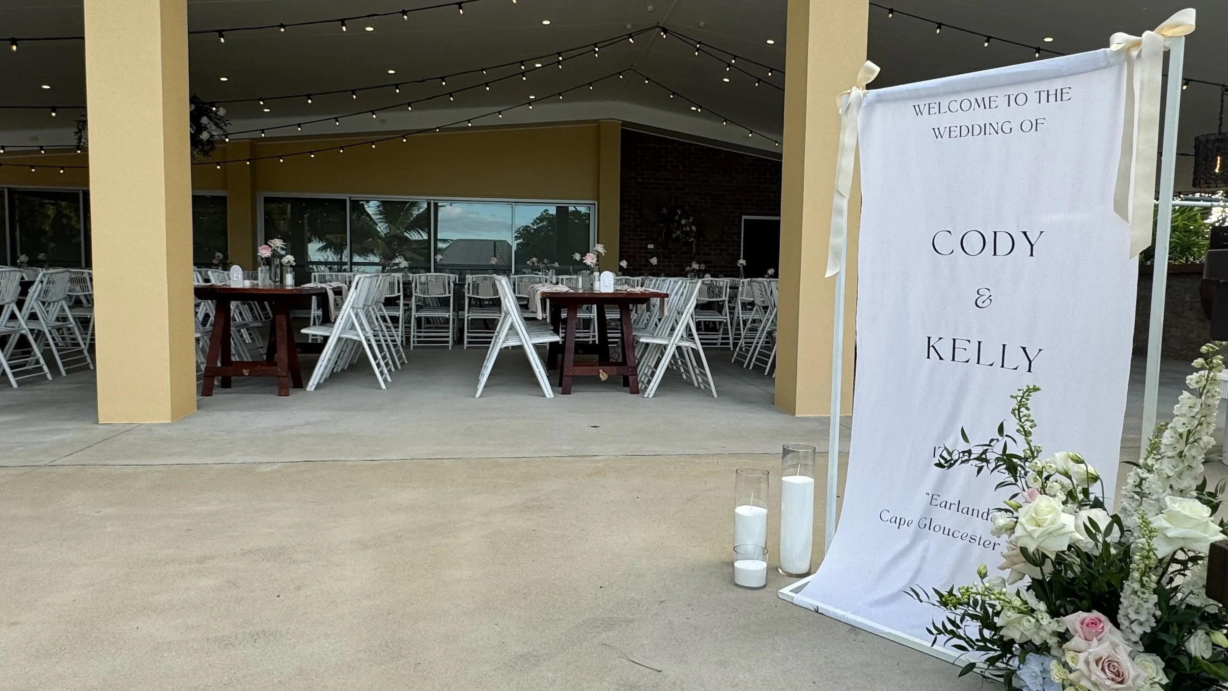 Outdoor wedding reception area with round tables and white chairs, decorated with floral centerpieces, under string lights. A welcome sign with wedding details stands to the right, adorned with flowers and candles.