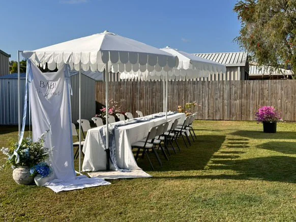 Outdoor wedding or event setup with a long table, white tablecloth, chairs, and a white canopy in a backyard with a wooden fence and potted flowers.