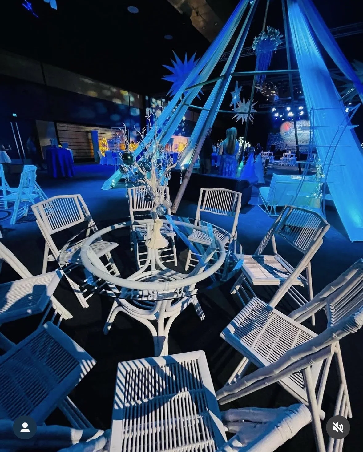 A decorated event space with a white round table surrounded by white chairs. The table has a centerpiece with ornaments and a small figurine. The background features large blue geometric star and snowflake decorations, with a blue and black color sch