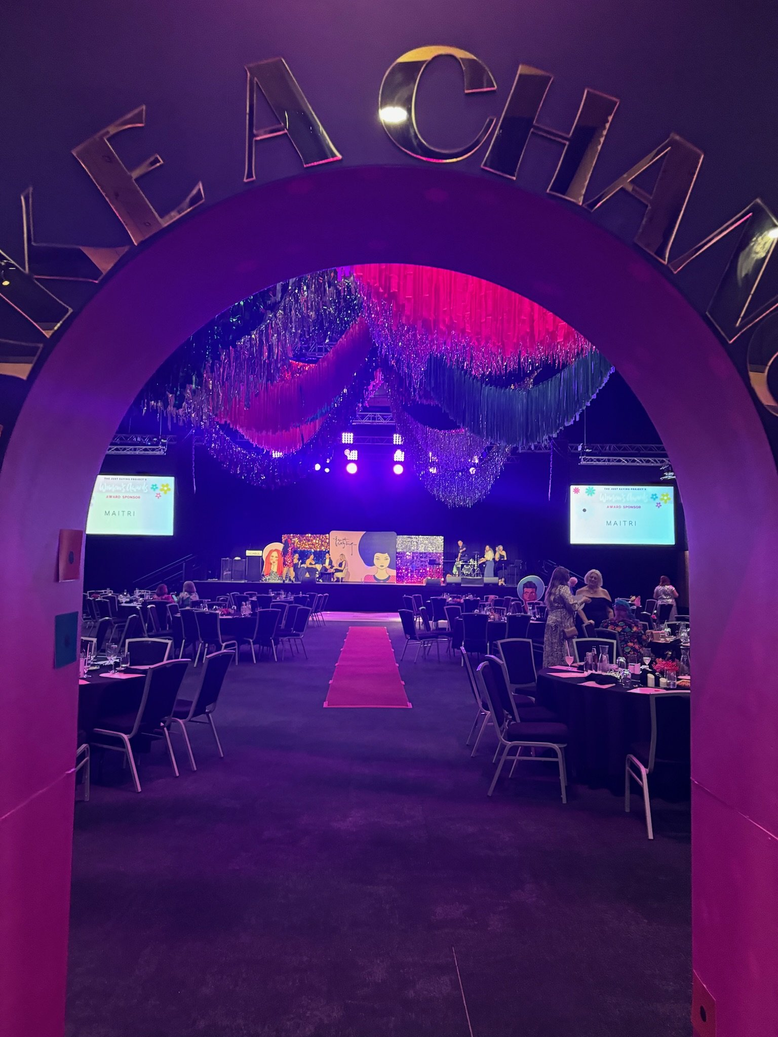 Empty banquet hall with a stage, decorated with colorful curtains and floral backdrop, set up for an event with round tables and chairs.