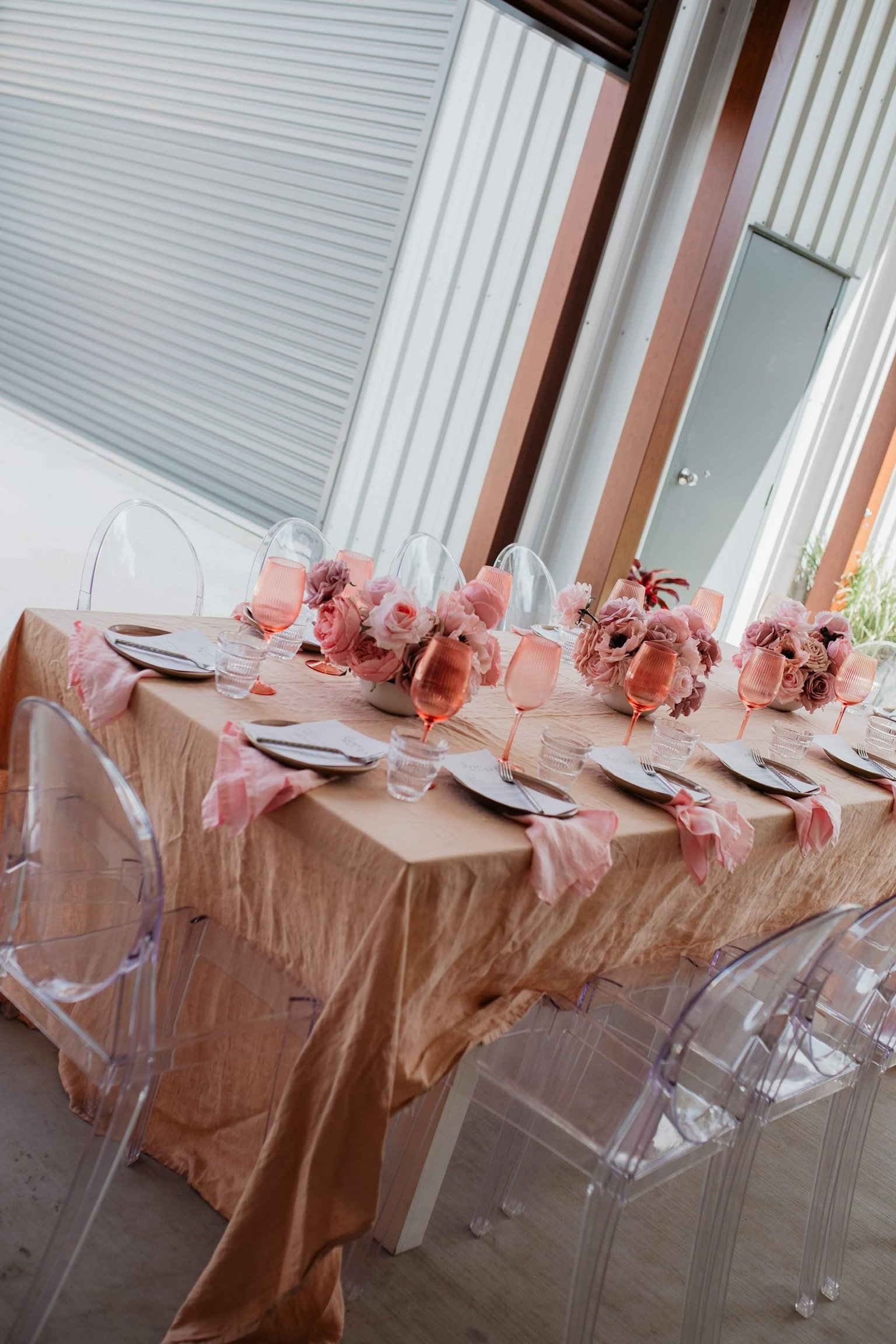 A rectangular dining table set for a formal event, decorated with a peach-colored tablecloth, pink floral centerpieces, pink-colored glasses, and place settings with forks, knives, and folded napkins. Transparent ghost chairs surround the table. The 
