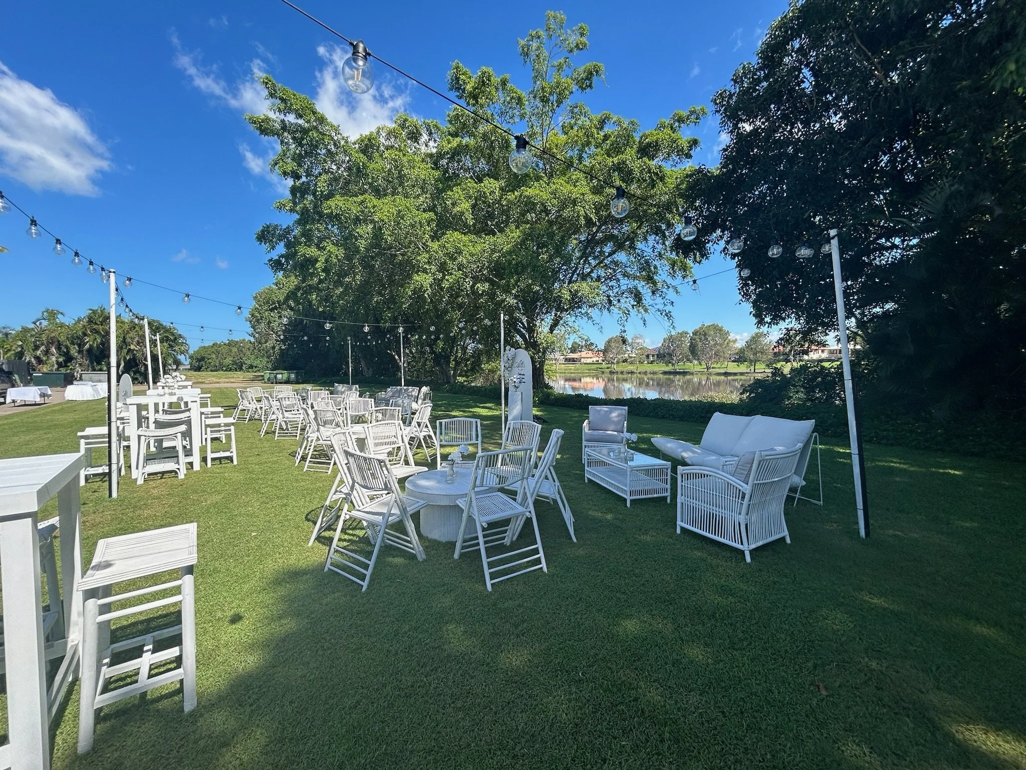 Outdoor event setup with white chairs and tables on a grassy area, string lights overhead, near a body of water, surrounded by trees under a clear blue sky.