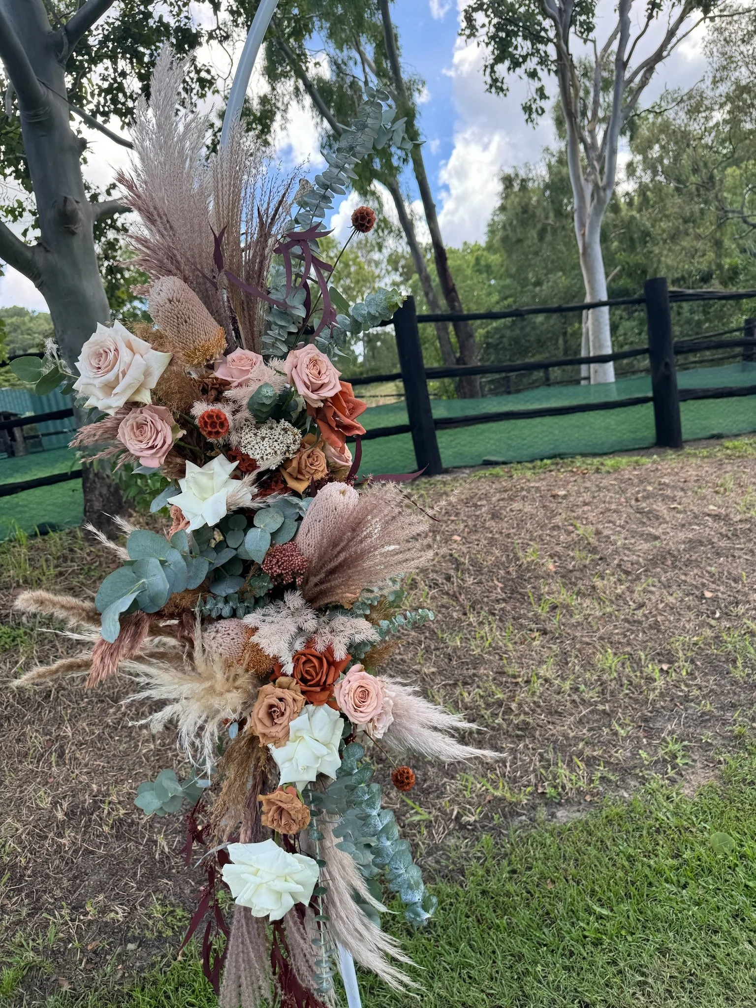 A floral arrangement with various dried and fresh flowers, including roses, eucalyptus, and pampas grass, attached to a fence outdoors on grass and dirt with trees and a cloudy sky in the background.
