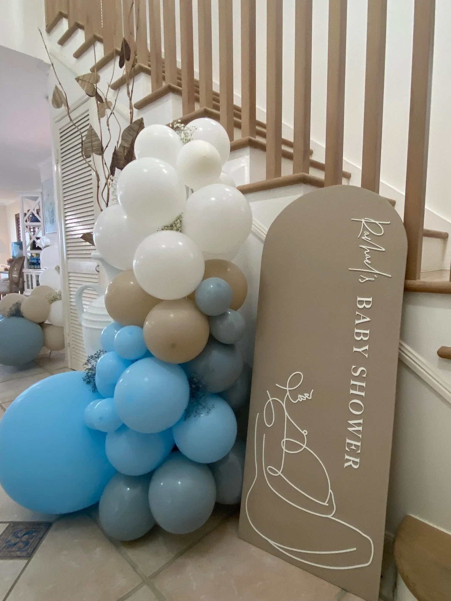 Decorative display of white, beige, and blue balloons next to a beige sign that reads 'RaRa's BABY SHOWER' with a line drawing of a baby wearing a hat.