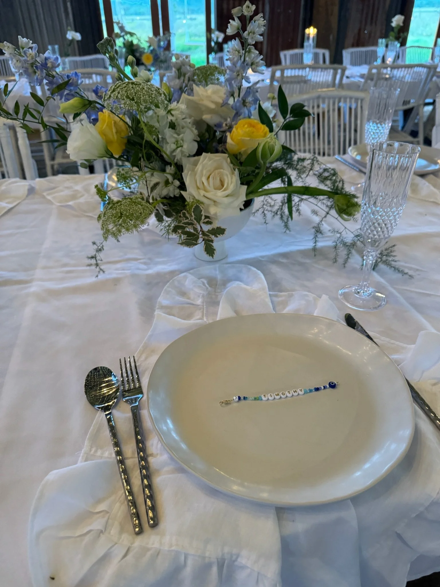 Table setting with a white plate, silver spoon and fork, clear glass goblet, white tablecloth, and a floral centerpiece with white, yellow, and blue flowers.