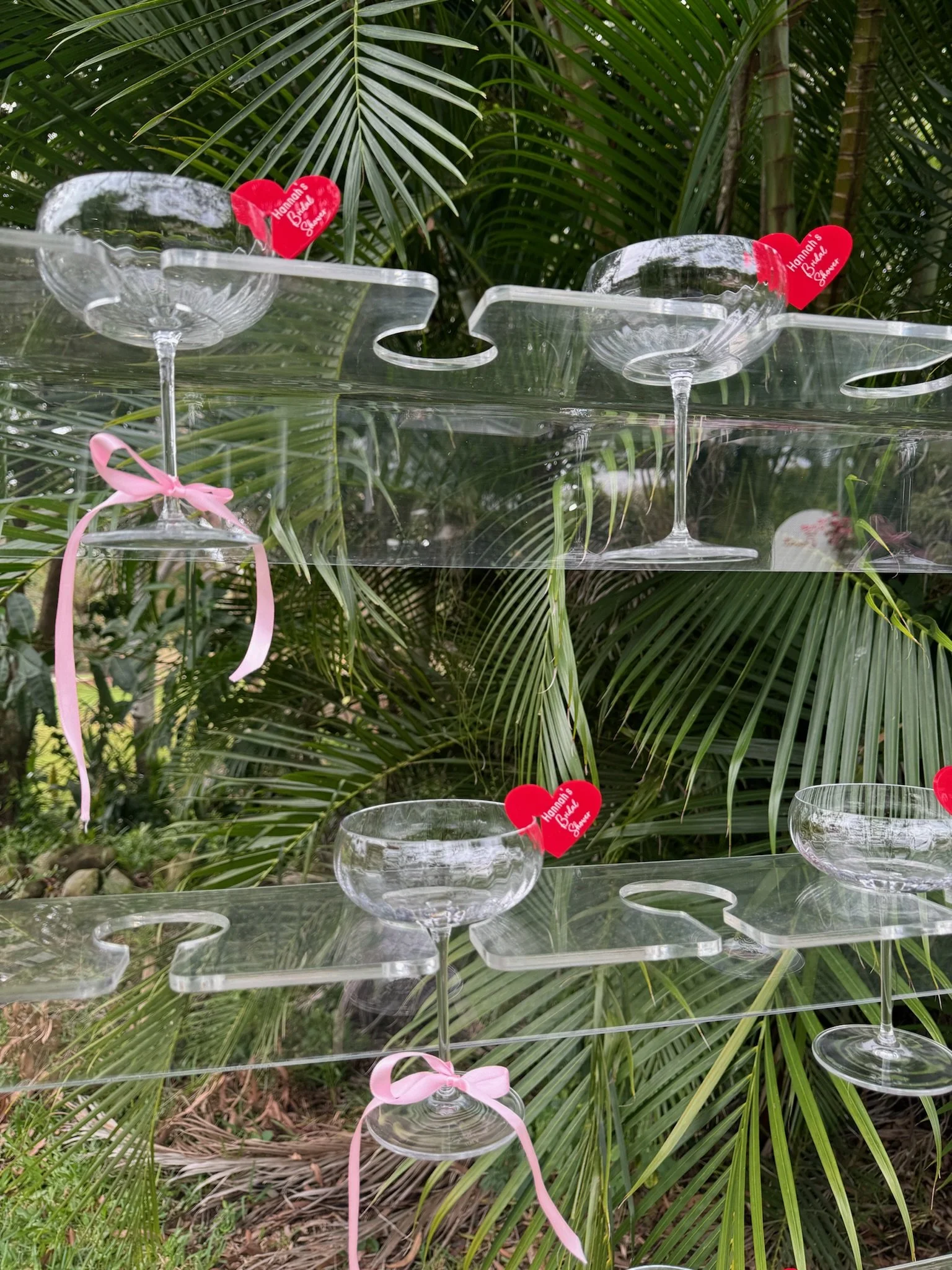 Empty glass dessert cups on a hanging acrylic shelf, decorated with pink ribbons and red heart-shaped tags, set against lush green palm leaves.