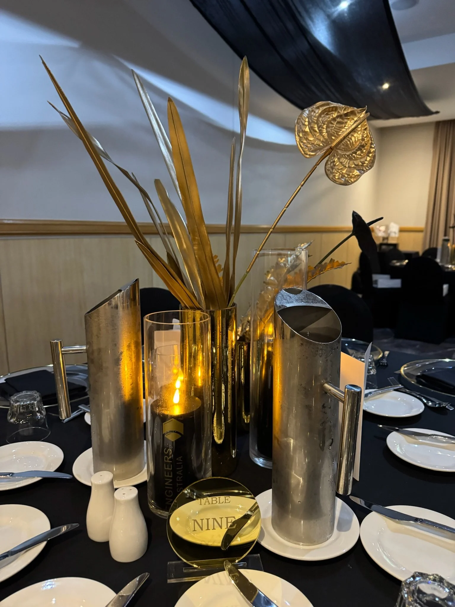 Elegant table centerpiece with gold and metallic silver leaves in tall vases, set on a black tablecloth with white plates and silverware, at a formal event.