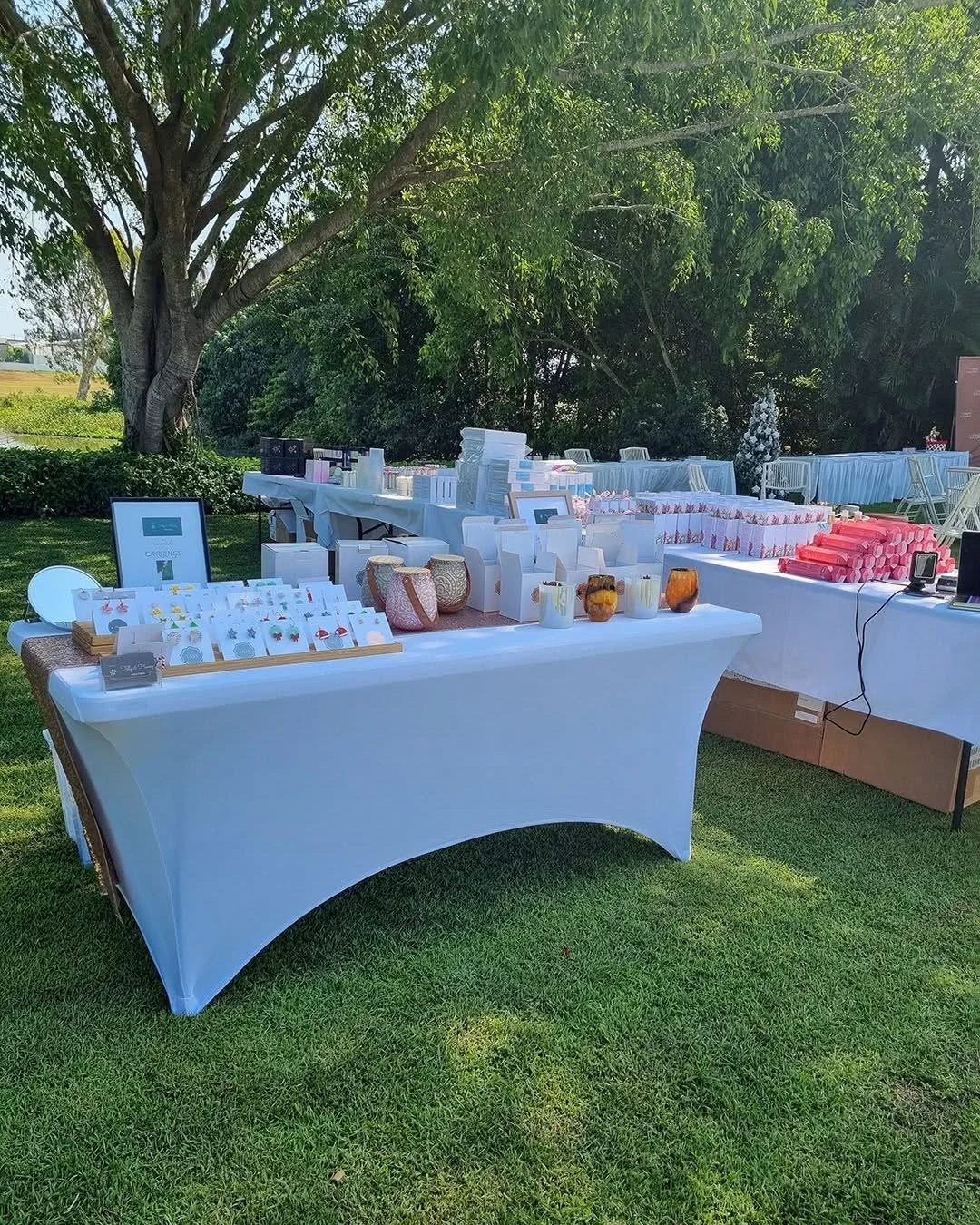 Outdoor view of a white table with various small decorative items and gifts, set up on a grassy area with trees in the background.