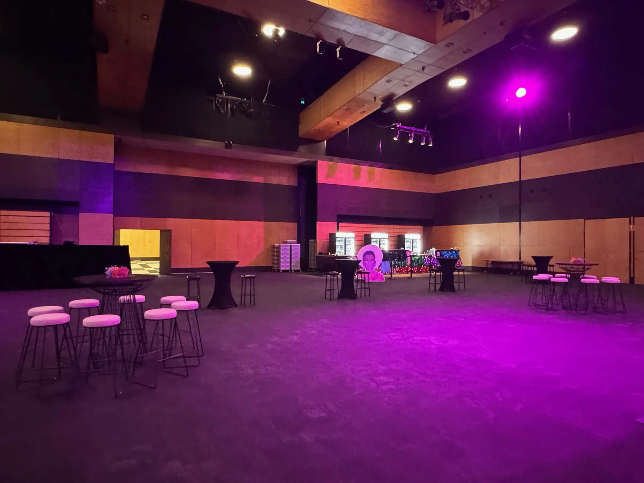 Empty event space decorated with purple and pink lights, high tables with stools, a bar area, and a photo booth with a portrait of a woman.