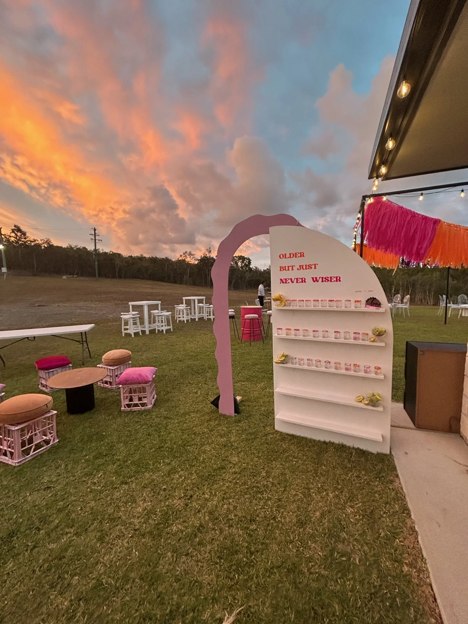 Outdoor event setup at sunset with white tables and pink cushioned chairs, a decorative white shelf with small containers, and a pink and orange feathered banner, under a colorful sky.