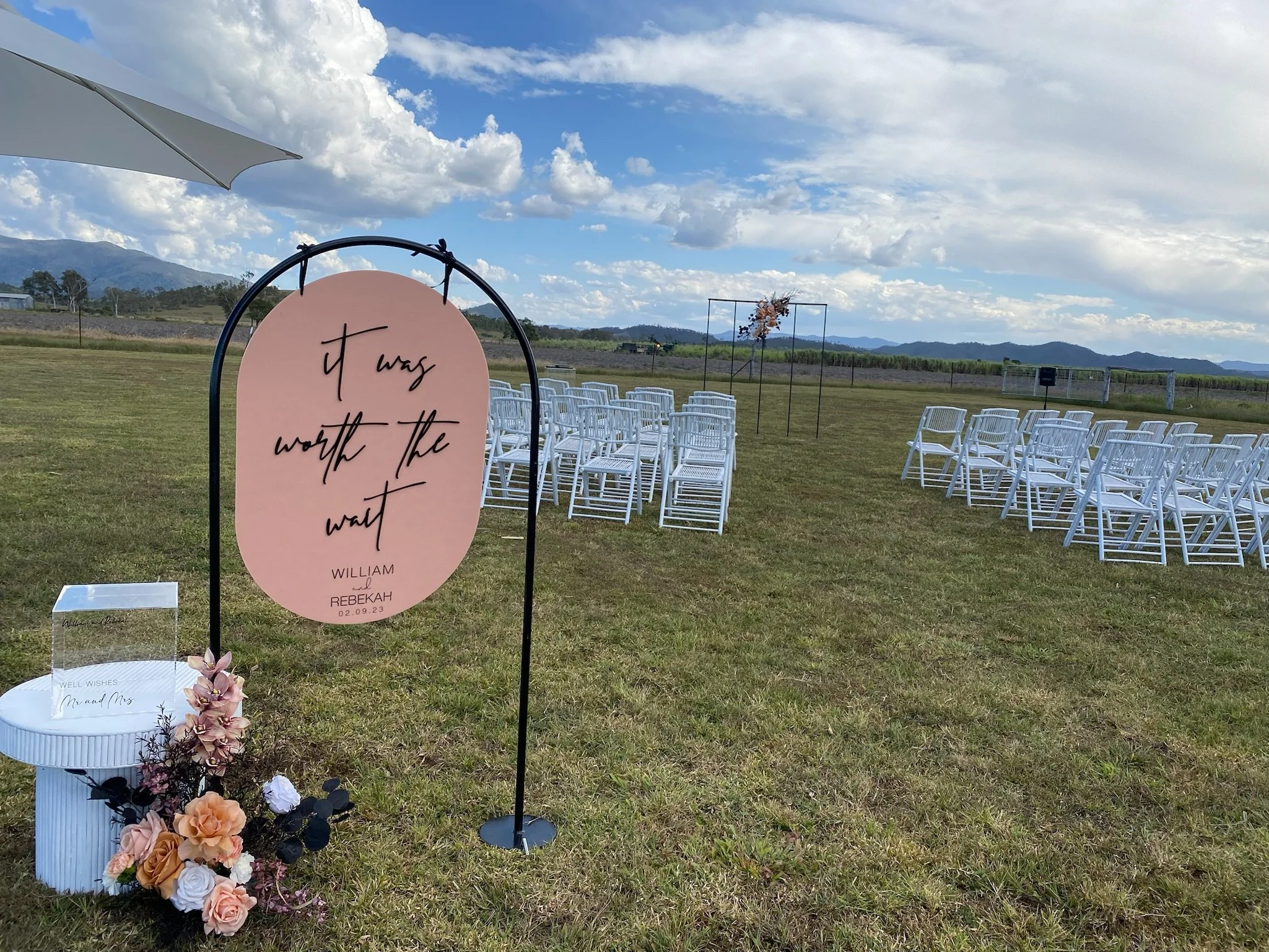 Outdoor wedding setup with white chairs arranged in rows on a grassy field, a pink sign with black handwritten-style text that reads 'it was worth the wait, William and Rebekah 02.09.23,' a white table with a floral arrangement, and a backdrop of mou