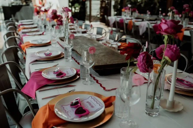 A long banquet table set for a special event with pink and orange napkins, floral centerpieces, glassware, and plates, in a decorated venue.