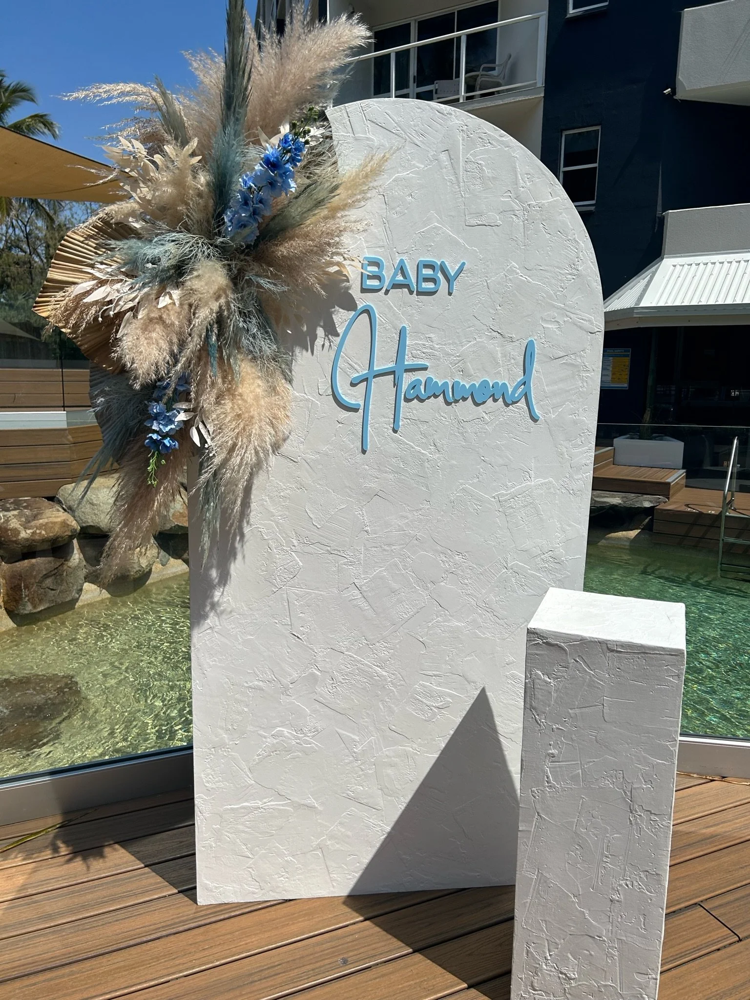 White textured sign with blue text reading 'Baby Hammond', decorated with a large arrangement of pampas grass, blue flowers, and dried leaves, situated on a wooden deck near a pool.