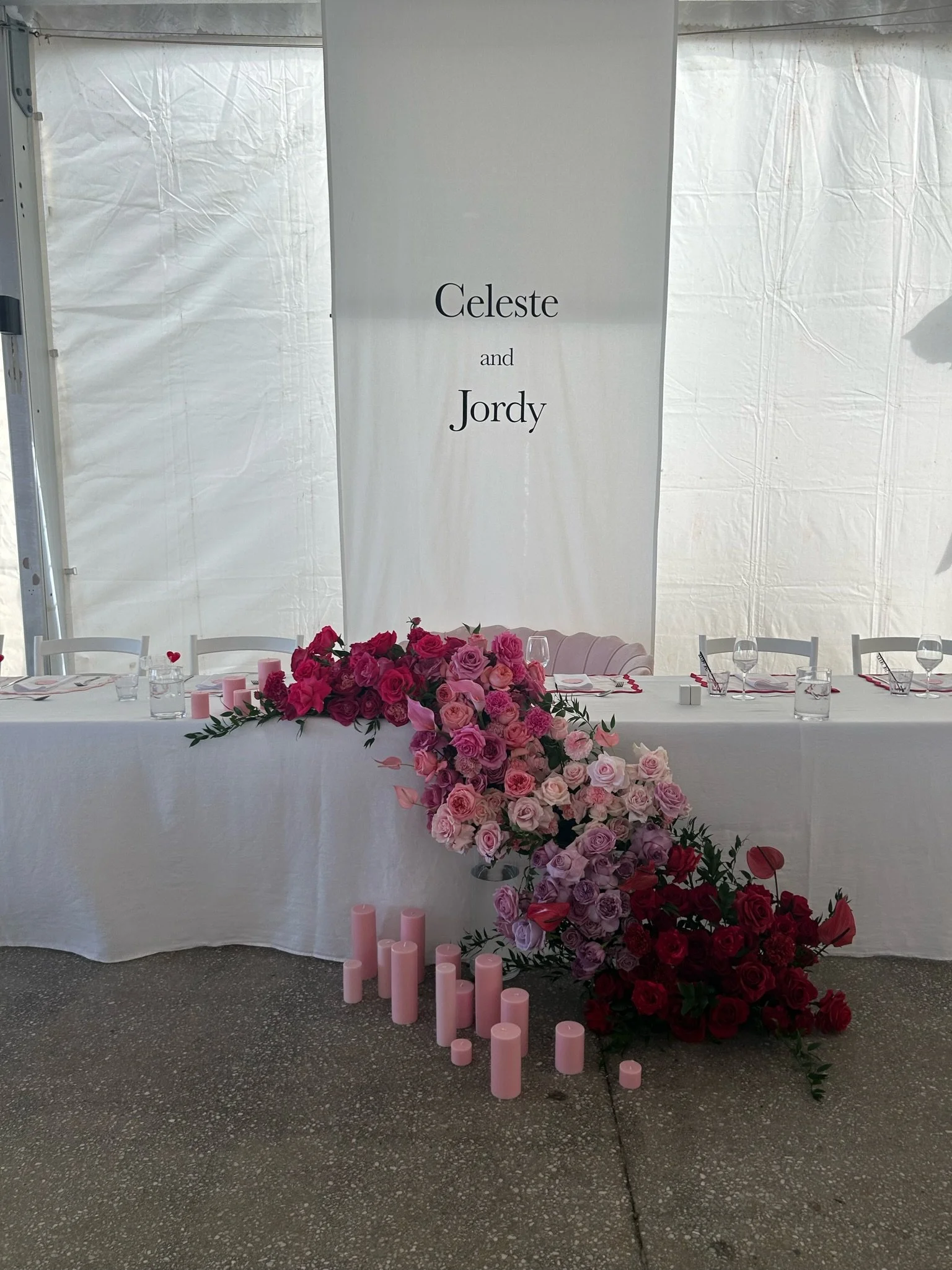 Wedding cake table decorated with pink and red roses, pink candles, and a white tablecloth, with a banner behind it reading 'Celeste and Jordy.'