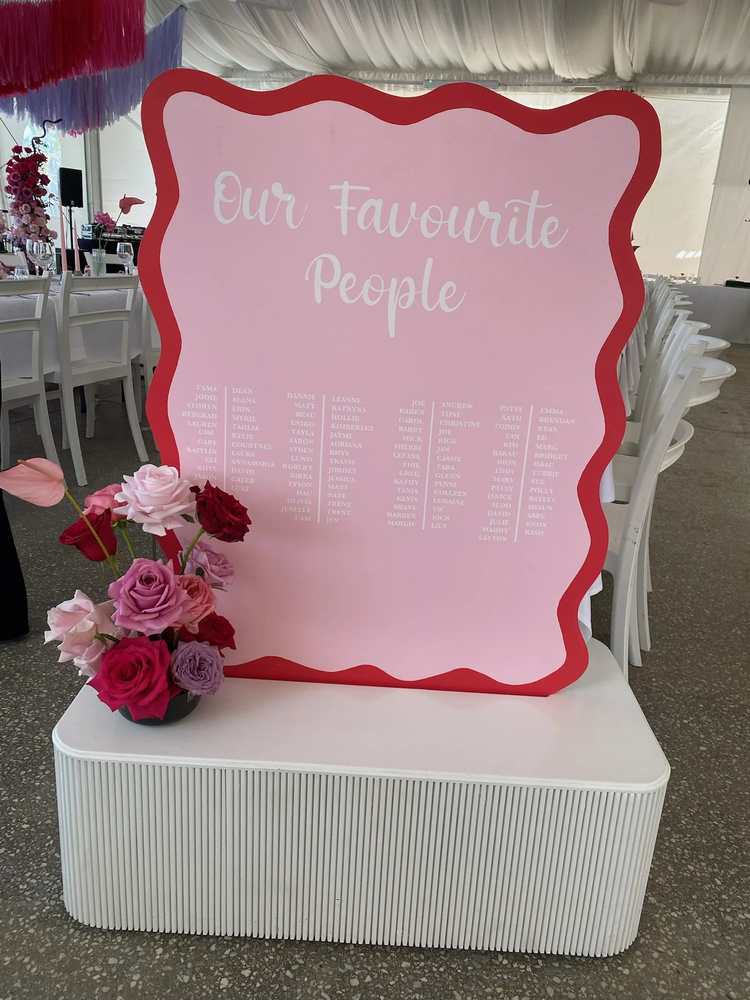 A pink board with red wavy border that reads 'Our Favourite People' and lists names, placed on a white table with a vase of pink, purple, and red roses. The background shows a decorated event tent with white chairs and tables.