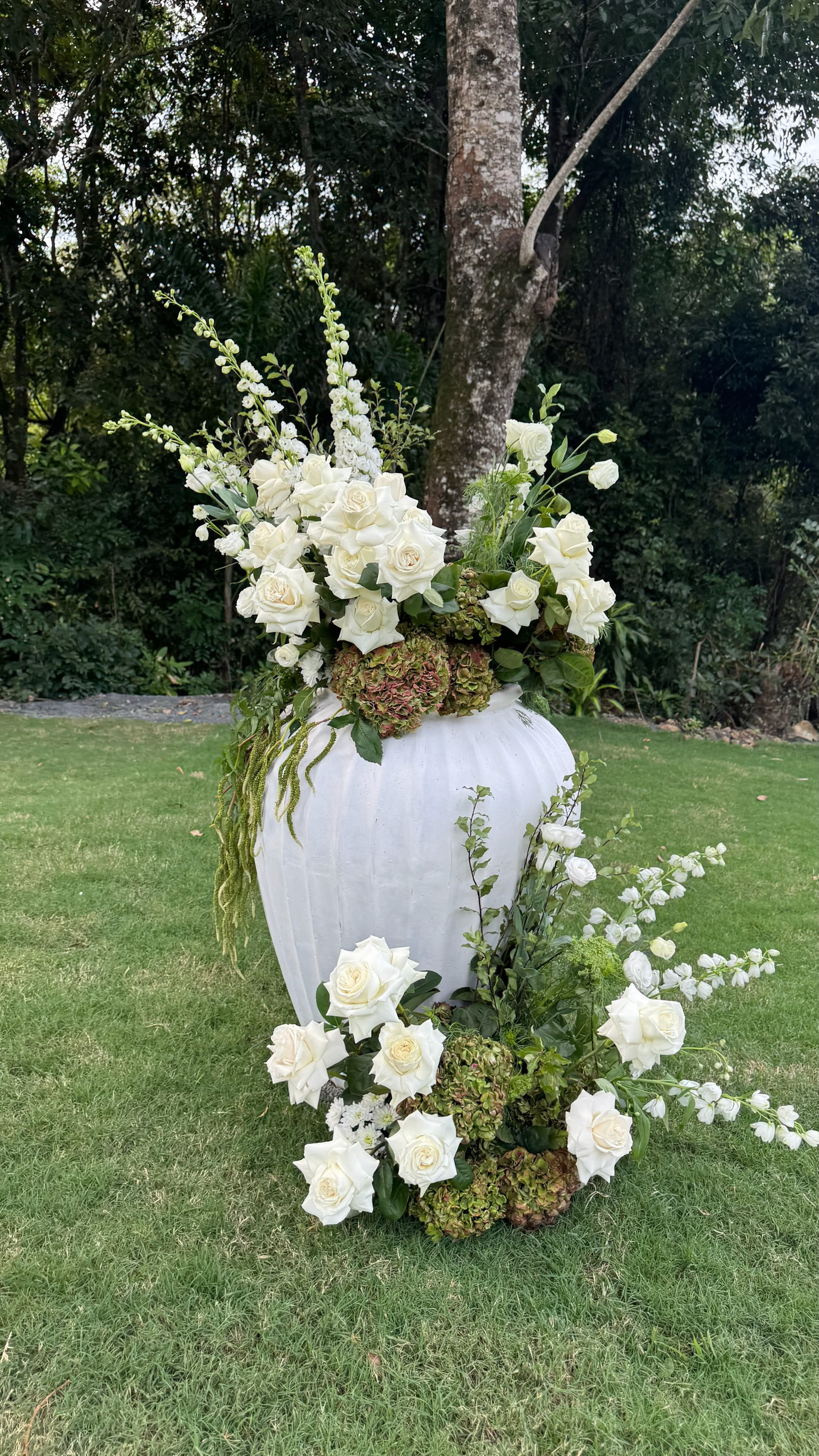 A large white vase filled with white roses, hydrangeas, and greenery, placed on the grass in a garden with trees in the background.