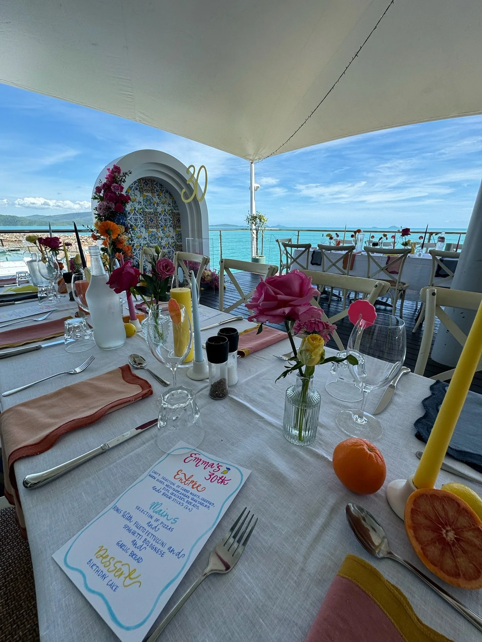 Decorated table set for a celebration on a balcony overlooking the ocean, with pink flowers, a menu, and a 30th birthday sign in the background.