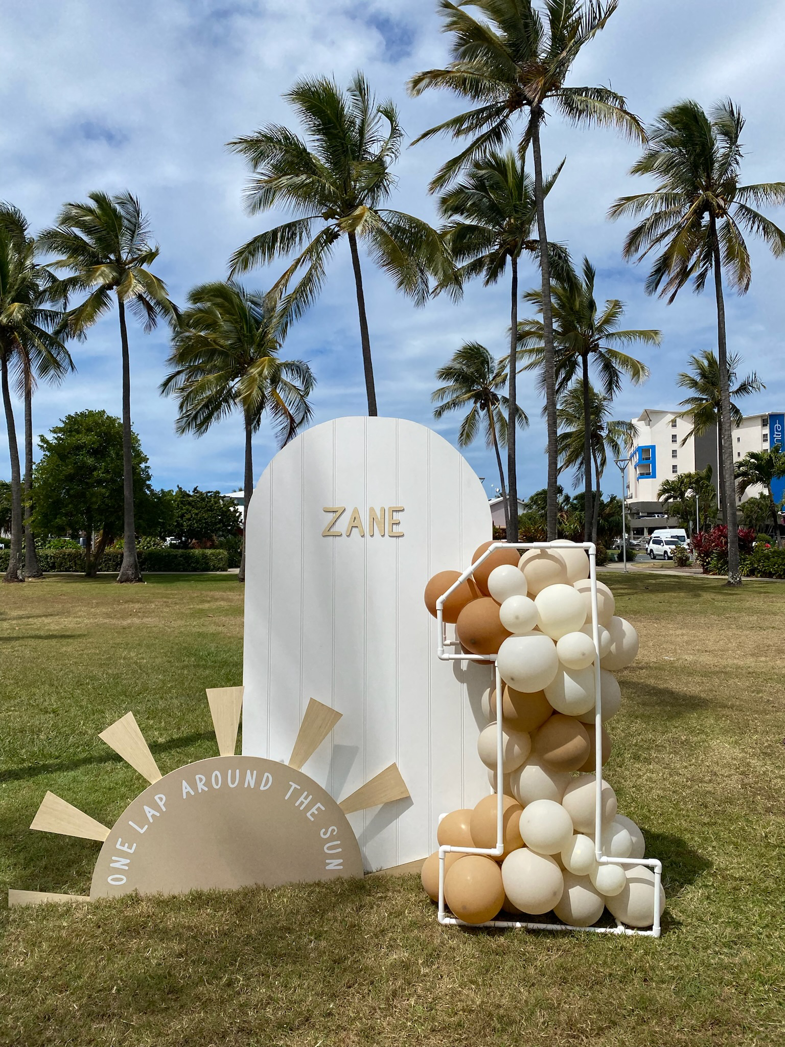 Decorative display outdoors with palm trees, a white door-like backdrop with 'ZANE' written on it, a semi-circular sign that says 'ONE LAP AROUND THE SUN,' and a cluster of tan, cream, and white balloons arranged in a metal frame.