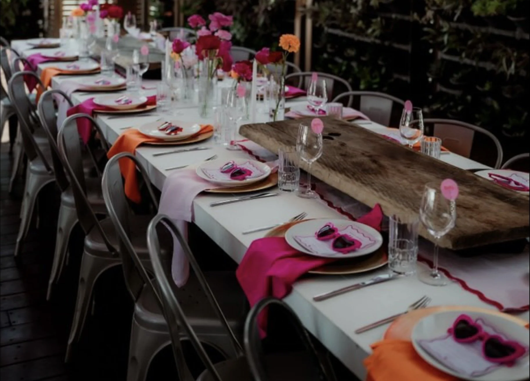 A long banquet table decorated with pink, orange, and red flowers in glass bottles, set with white plates, pink and orange napkins, silverware, wine glasses, and small pink tags or place cards, in an outdoor setting with greenery.