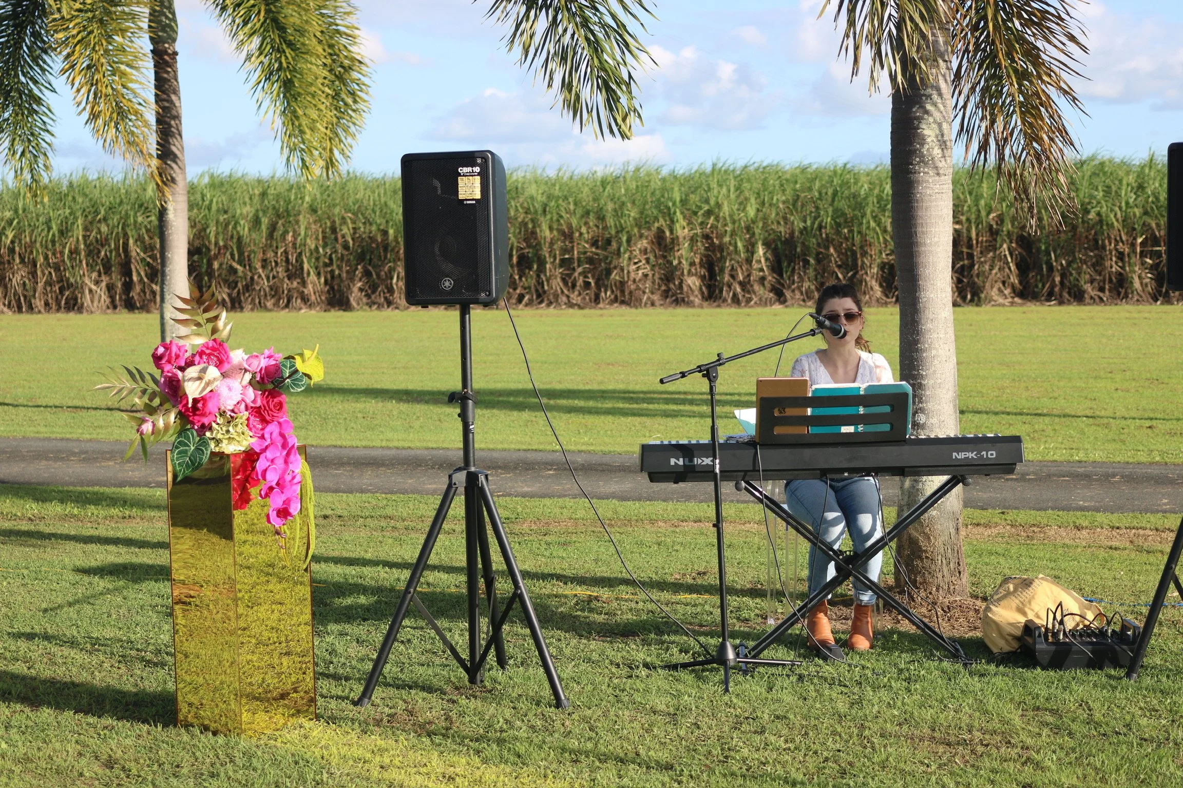 A woman wearing sunglasses singing or speaking into a microphone at an outdoor event, playing a keyboard, with speakers and a floral arrangement nearby, set against grassy fields and palm trees.
