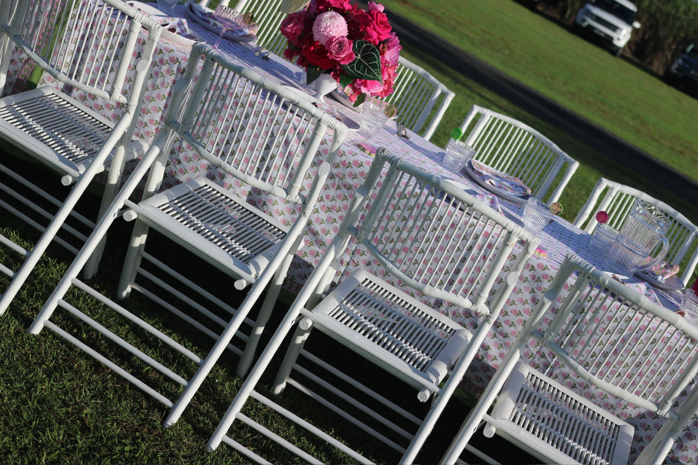 Outdoor dining table set with white chairs, floral tablecloth, pink floral centerpiece, plates, glasses, and napkins on a grassy area.