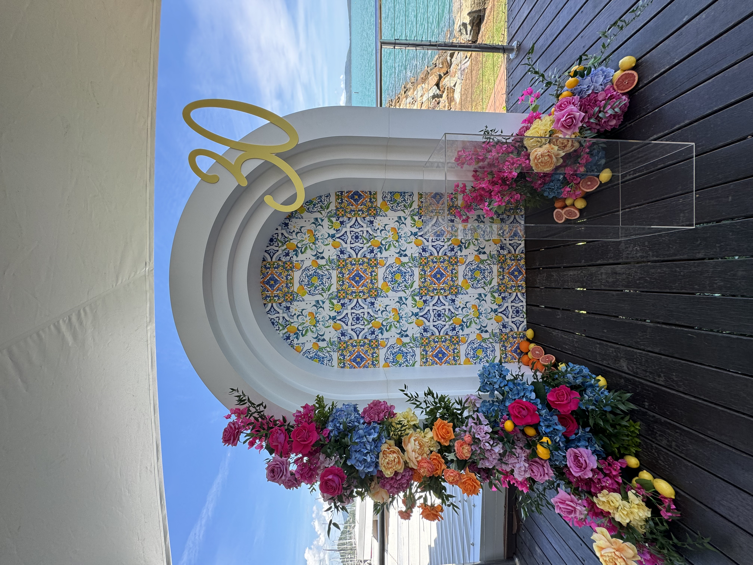 Decorative floral arrangement with pink, purple, blue, yellow, and orange flowers on dark wooden deck in front of a white arch with colorful patterned tiles, overlooking a waterfront with a boat in the distance.