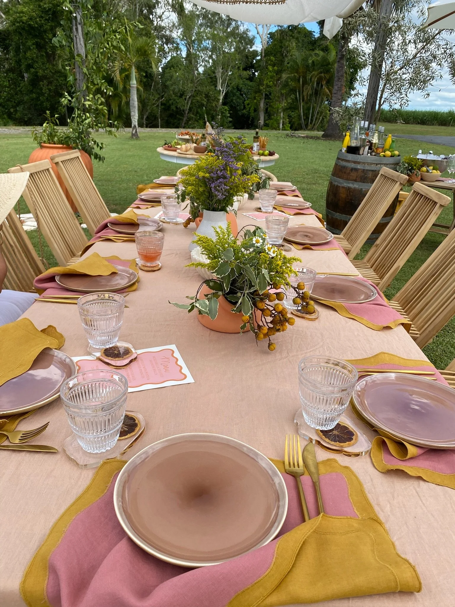 Outdoor table set for a gathering with a pink tablecloth, gold and pink place settings, and floral centerpieces. In the background, there are tables with food and drinks, and the setting is surrounded by green trees and a grassy field.