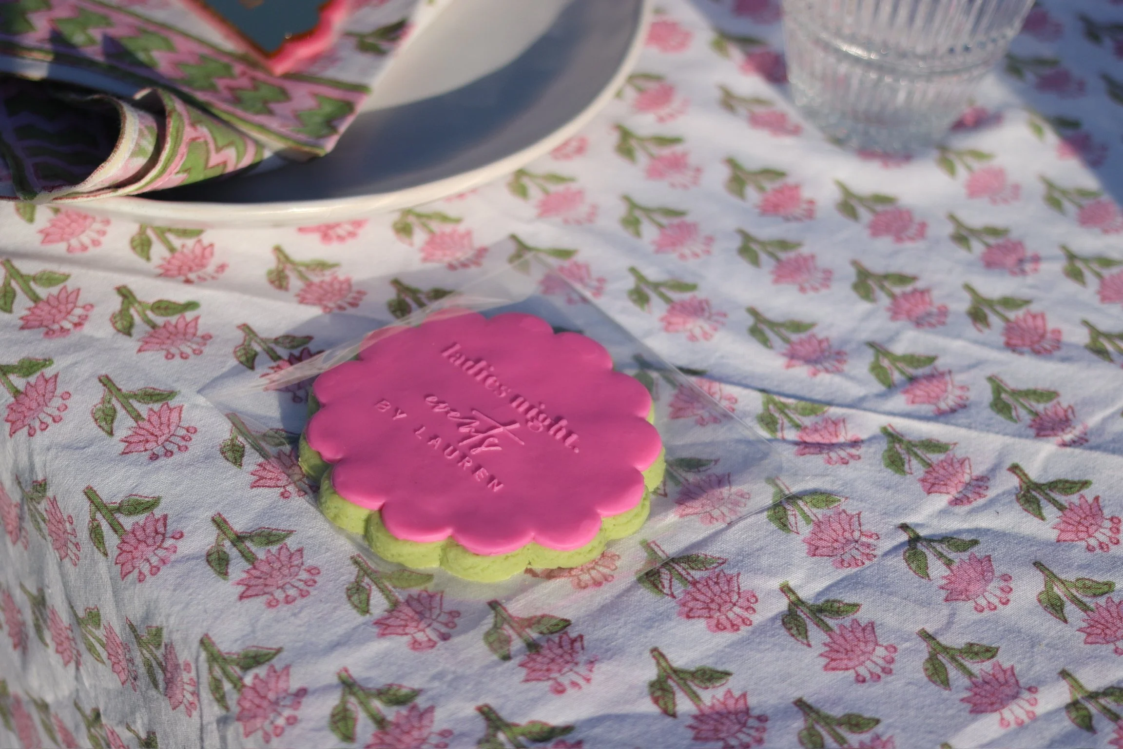 A pink and green cookie with writing on it, placed on a floral tablecloth, beside a glass of water and a white plate with a patterned napkin on top.